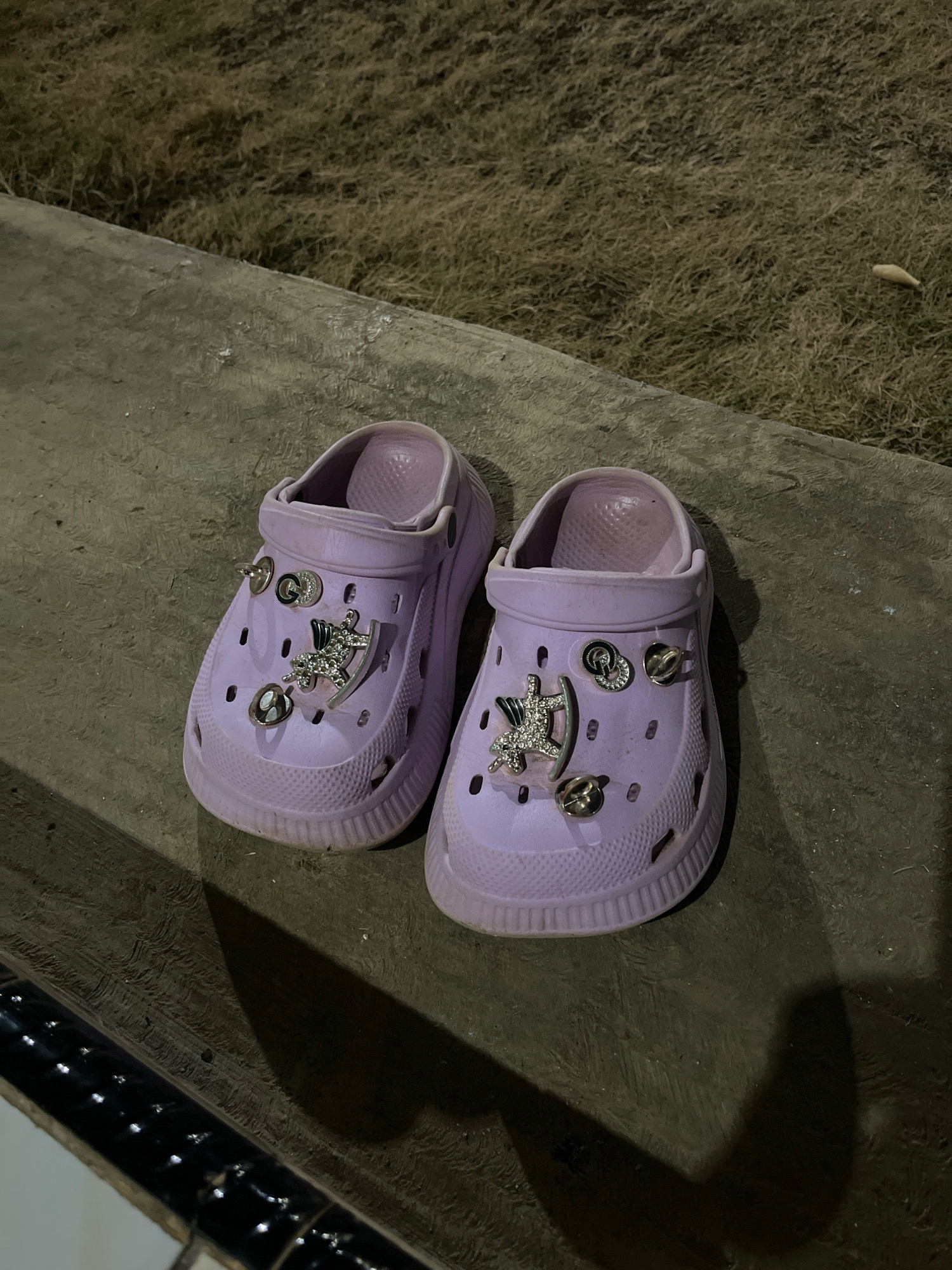 A pair of light purple Crocs, adorned with various metallic charms including a horseshoe, a rocking horse, and round decorative pieces, are placed side-by-side on a textured concrete surface. Behind the Crocs, dry, straw-like grass is visible. The scene appears to be outdoors, possibly in a garden or yard area. The lighting suggests it is evening or night, with shadows cast on the concrete. There is no indication of the specific location within Kebumen, Indonesia.
