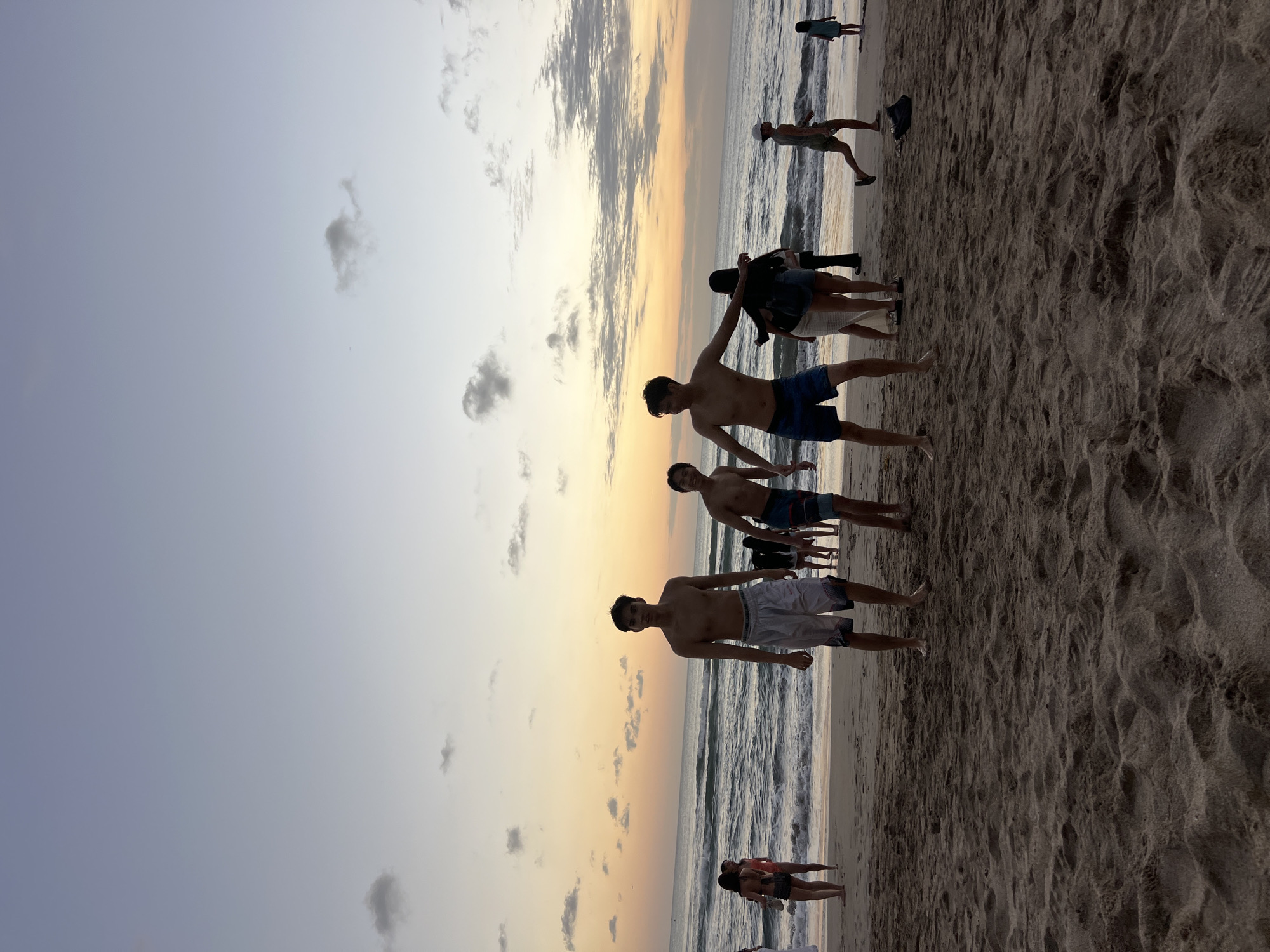The image depicts a beach scene at dusk, likely in Kuta, Indonesia, given the presence of a tropical beach environment. The sky is a gradient of soft blues and oranges, indicative of sunset, with scattered clouds. The ocean stretches to the horizon, with gentle waves breaking on the shore. The sandy beach is populated by several individuals, most of whom are young adults and teenagers.
In the foreground, three young men are standing on the sand, facing towards the camera. The man in the center is wearing patterned swim trunks and is shirtless. To his right, another shirtless young man is wearing blue swim trunks. To the left of the central figure, a third shirtless young man is wearing white swim trunks with dark patterns.
Further back, other beachgoers are visible. On the left side, a young woman in a swimsuit is standing near the water. To her right, several other people are seen walking or standing on the beach. In the middle ground, near the water's edge, a group of people are gathered. To the right, a young man wearing a cap and green shorts is walking away from the camera, and a young girl in a blue dress is standing closer to the water. The sand is textured with footprints and ripples.