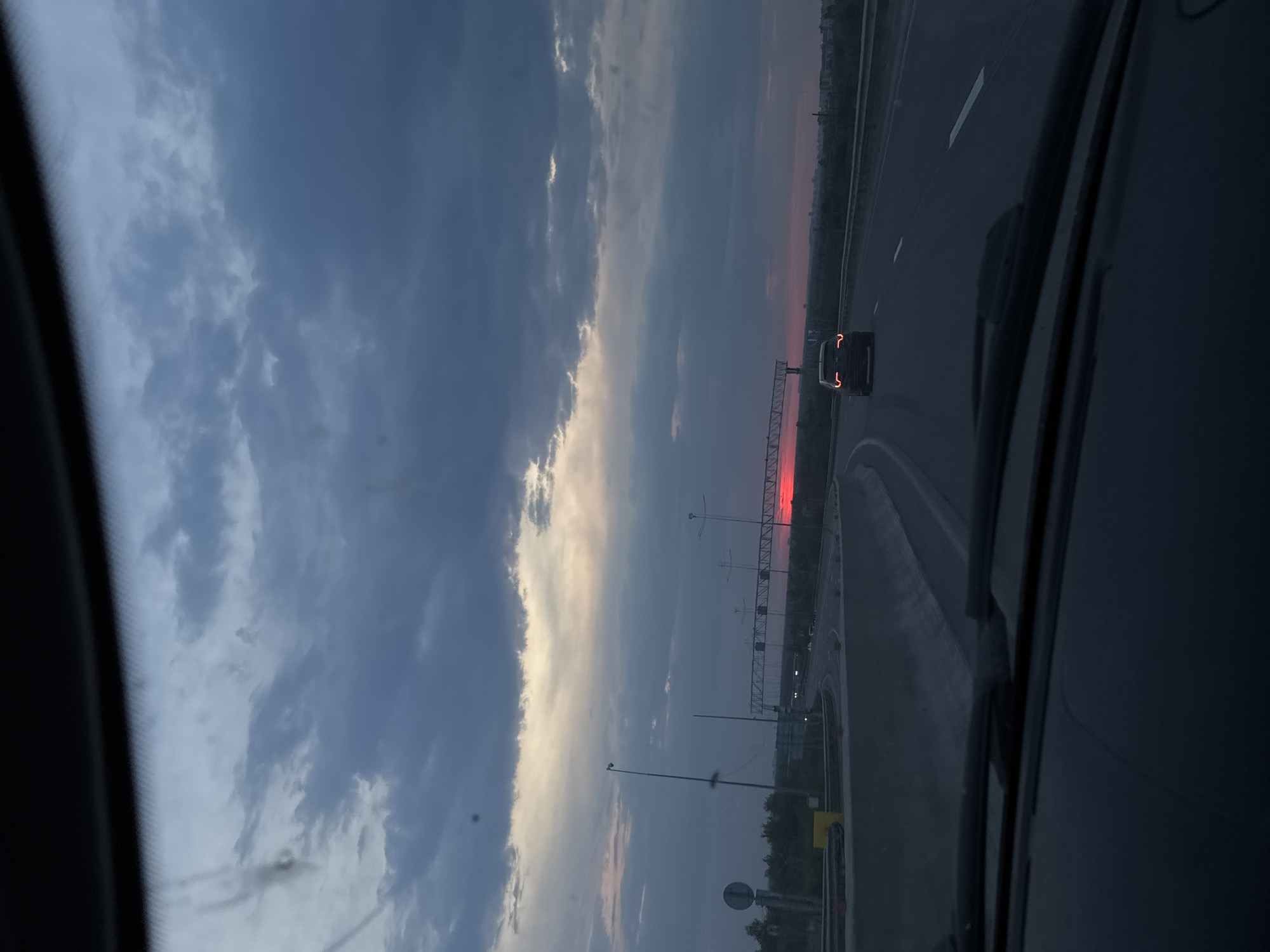 The image depicts a view from inside a vehicle, likely a car, traveling on a highway. To the left, a cloudy sky is visible with hues of blue and grey, tinged with light suggesting a sunrise or sunset. The highway stretches ahead, with a vehicle visible in the distance. Infrastructure like support beams, poles, and vegetation are along the side of the road. The image has a slightly rotated orientation. Given the location in Кондратово, Russia, it is likely the outskirts of the city, traveling along one of the major highways connecting it to surrounding areas.