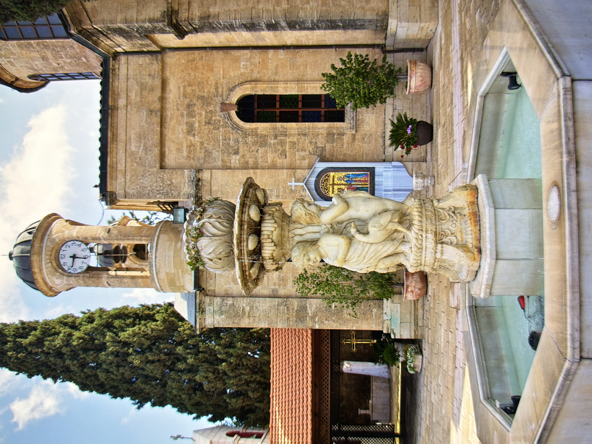 This is a medium shot of a fountain in front of a light brown stone building with a domed bell tower. The bell tower has a clock face with Roman numerals. To the left of the fountain is a tall evergreen tree and a building with a red tiled roof. The fountain itself is made of stone and features sculptures of cherubs and a bird. The water in the fountain is clear and has a slight ripple. There are potted plants and a mosaic icon in the background. The ground is paved with square stones. The sky is blue with some white clouds.