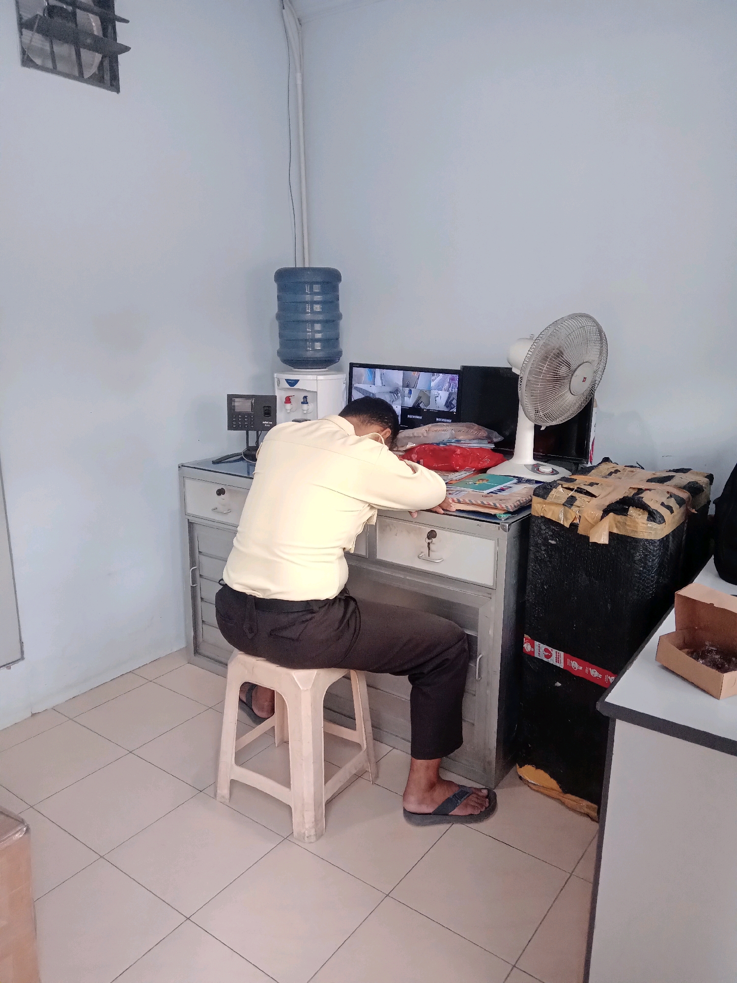 A person wearing a light-colored shirt and dark pants is asleep at a desk in what appears to be a security or office room in Surabaya, Indonesia. The person is leaning their head on their arms, which are resting on the desk. In front of the person is a computer monitor displaying multiple security camera feeds. To the left of the monitor is a water cooler with a large blue water jug. Various office supplies, including books and papers, are on the desk. A white oscillating fan is positioned behind the monitor. To the right of the desk, there is a large black object covered in packing tape and a small open cardboard box containing dark items, possibly food. The room has light blue walls and a tiled floor.