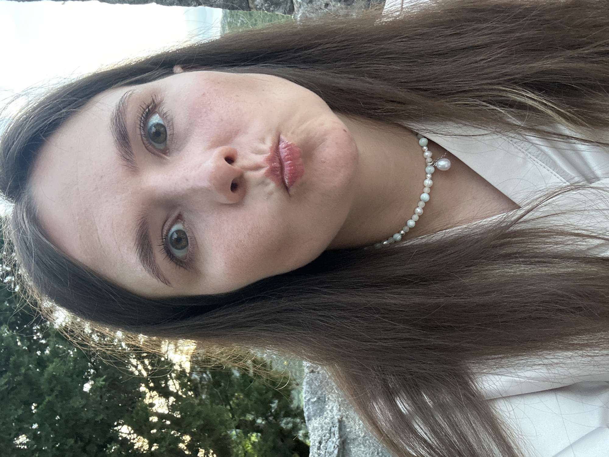 A young woman with long, brown hair and light skin is captured in a close-up, selfie-style portrait. She is outdoors, with a blurred background of green trees, a stone wall, and a bright sky. Her eyes are wide open, and her lips are pursed in a playful expression. She is wearing a white collared shirt and a pearl necklace with a single large pearl pendant. The lighting suggests it is late afternoon or early evening.