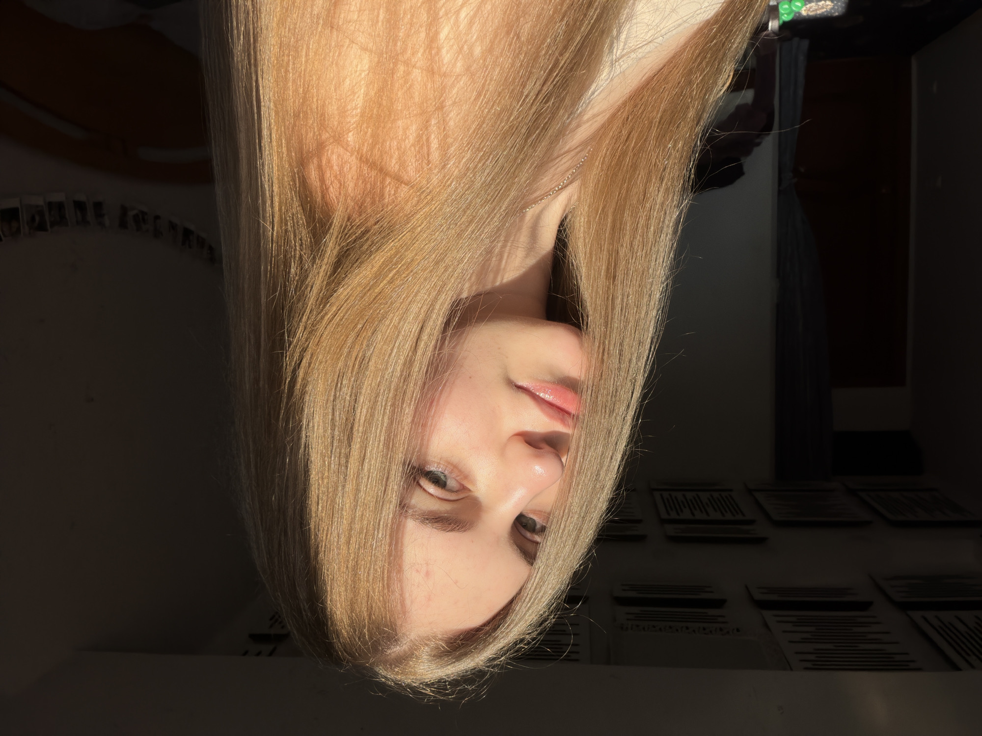 The media file features a close-up, upside-down shot of a young woman's face, illuminated by warm, natural sunlight. Her fair skin is highlighted, with a slight sheen on her nose and cheeks. She has light blonde hair that is long and straight, framing her face and partially obscuring her features. Her eyes are a light brown or hazel color, and her lips are slightly parted, with a glossy finish. A delicate silver necklace is visible around her neck.
The background is dark and out of focus, but suggests an indoor setting. On the left side, a row of small, framed photographs is dimly visible, arranged in an arc against a shadowed wall. The right side of the background is even darker, with hints of furniture or room dividers. Scattered across the lower portion of the frame are rows of dark objects that appear to be flat, rectangular items with linear patterns, possibly records or stacked books.
The lighting strongly suggests that the photo was taken during golden hour, with the low-angled sunlight creating a warm, inviting atmosphere. The overall mood is serene and contemplative, with the natural light accentuating the subject's features. The presence of the photographs hints at a personal space, likely a bedroom or living area in a home. The location context provided is Dnipro, Ukraine. No discernible text is visible in the image.