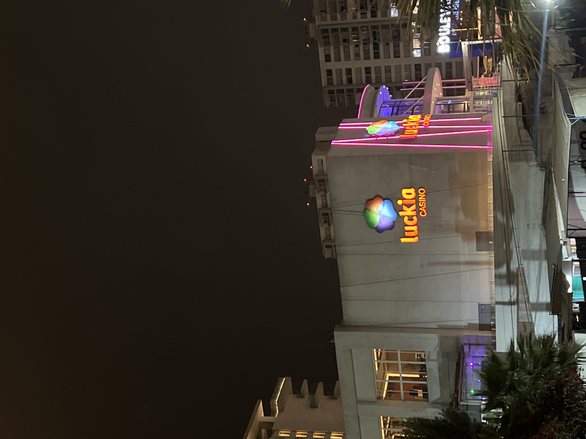 The image shows the Luckia Casino in Arica, Chile, at night. The casino building is brightly lit with the Luckia logo visible, and there are pink neon lights running along the top of the building. A tall building is behind the casino. The sky is dark. Palm trees are in the foreground.