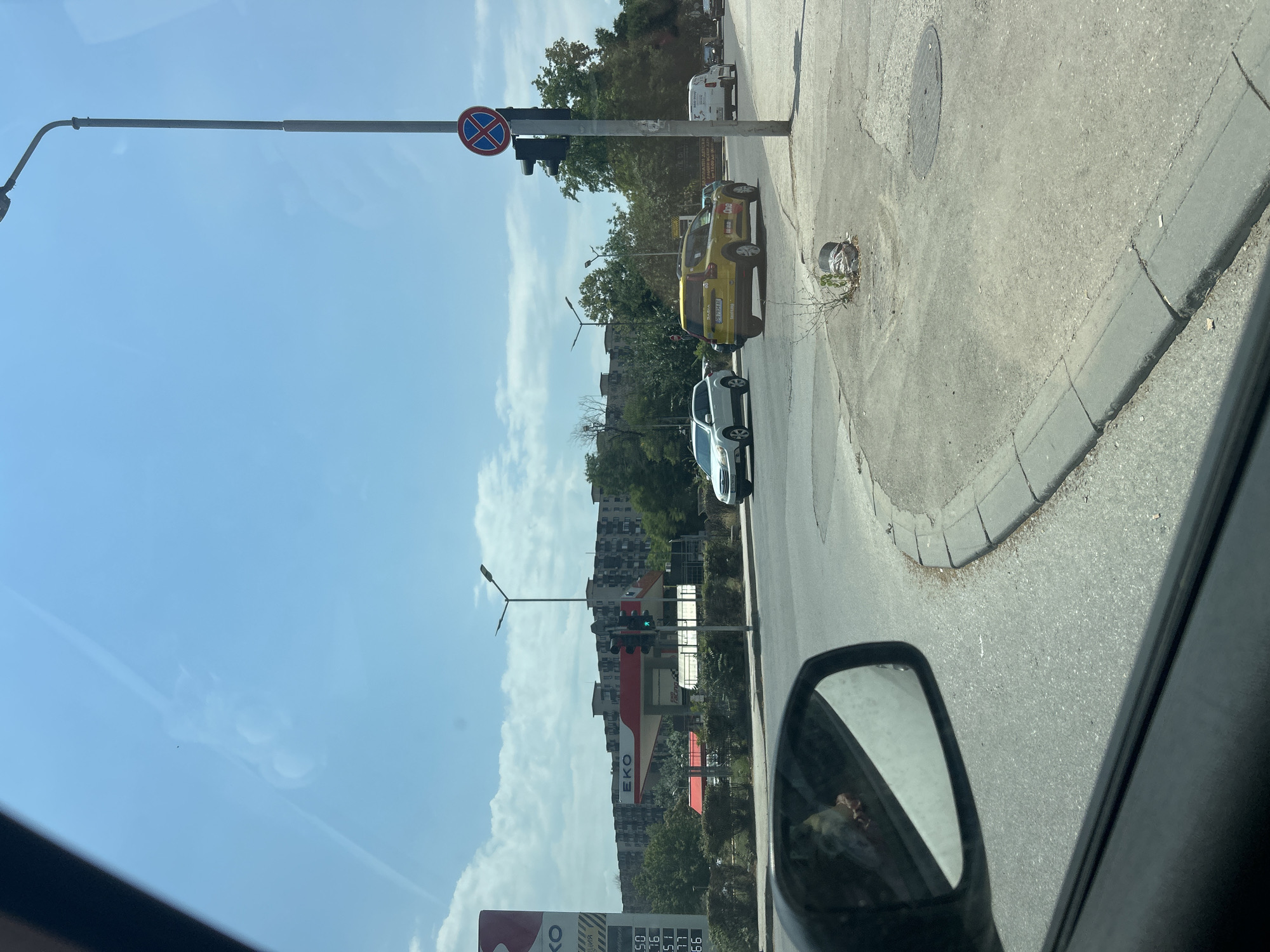 This media file depicts a street scene in Plovdiv, Bulgaria, likely on a sunny day with a clear blue sky and scattered clouds. The perspective is from inside a vehicle, looking out the side window and into the driver's side mirror.
In the foreground, the driver's side mirror is visible, reflecting a blurry image of what appears to be a person and a car interior. The main scene shows a street with asphalt pavement and a concrete curb. A lamppost with a "no stopping" traffic sign is prominent on the left side of the road. Further down the street, a yellow taxi, possibly a Dacia, is parked. A white car is also visible.
Beyond the immediate roadside, there are trees and buildings, including apartment blocks. A traffic light is visible and appears to be green, indicating that traffic is flowing. In the background, a red and white structure with "EKO" written on it suggests a gas station.
The image captures an everyday urban scene, with vehicles and street infrastructure. The time of day appears to be daytime, given the bright sunlight and clear sky. The overall impression is one of a typical day in a city. The visible text includes the "no stopping" sign and the "EKO" logo on the gas station.