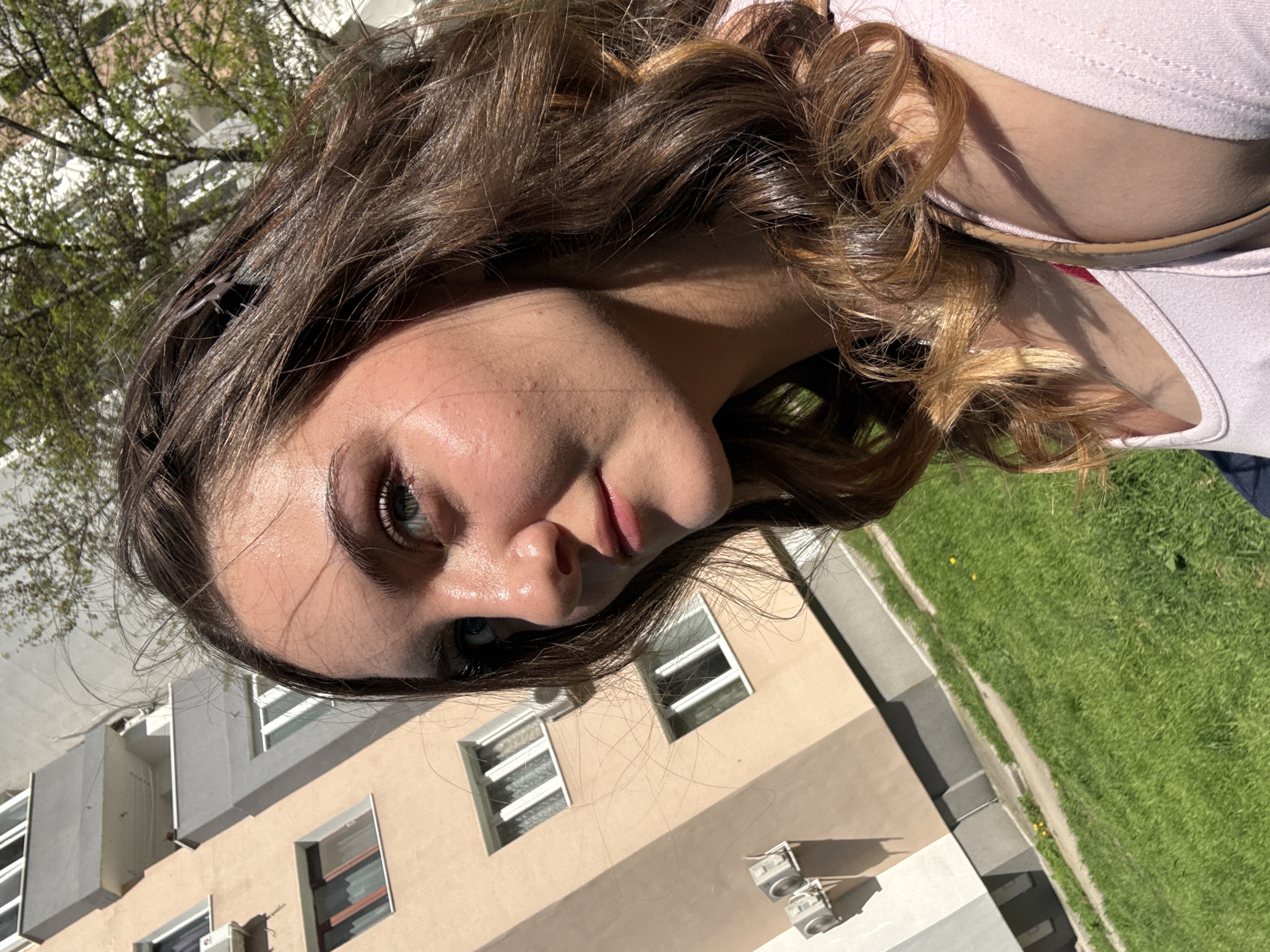 An outdoor, eye-level portrait-style shot captures a young woman, likely in her late teens or early twenties, against an urban backdrop in what appears to be Sofia, Bulgaria. The woman is positioned in the foreground, looking subtly towards the camera. Her long, wavy brown hair, featuring lighter, possibly highlighted strands, appears shiny and frames her face. She has striking blue eyes and her skin looks somewhat dewy under the bright sunlight. She is wearing a light pink top, partially visible on her right side.
The background reveals a multi-story apartment building with light beige and gray sections and numerous rectangular windows. Several external air conditioning units are mounted on a lower part of the building. To the building's right, a vibrant patch of green grass, possibly a courtyard or communal area, is visible, dotted with a few small yellow flowers. Above the building, the bare branches of a tree with emerging green leaves suggest the season is spring. The strong natural sunlight casts distinct shadows, indicating it is daytime. There is no visible text in the image.