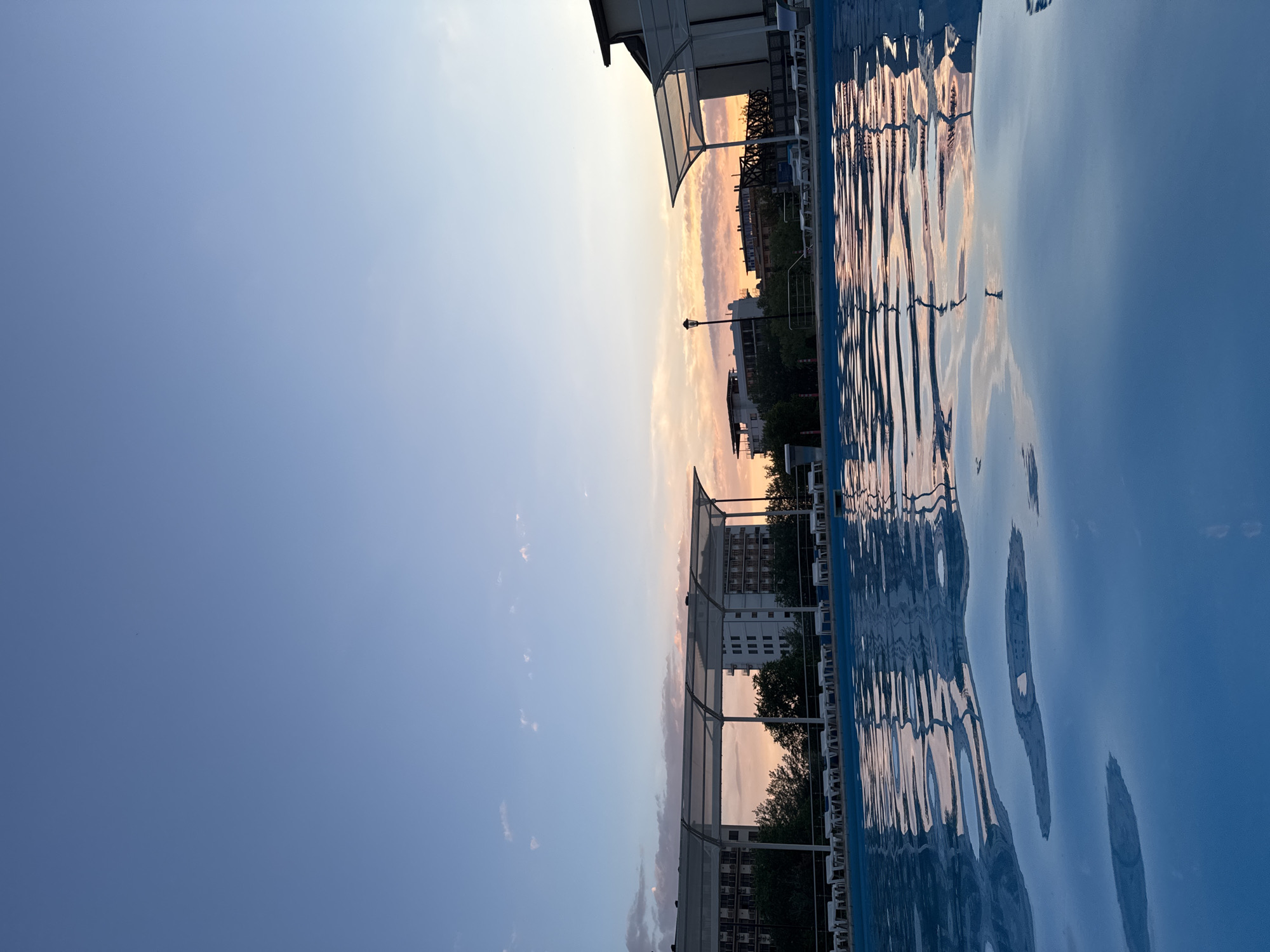The image depicts a swimming pool in Vityazevo, Russia, at dusk. The pool's surface reflects the colorful sky, which is a gradient of blues, pinks, and oranges, with a few scattered clouds. In the background, several buildings are visible, including multi-story hotels and smaller structures. Some of these buildings have white awnings extending over outdoor seating areas. White lounge chairs are neatly arranged along the edge of the pool. Trees and greenery separate the pool area from the buildings. A streetlamp is also visible in the distance. The scene is tranquil and appears to be in a resort or hotel setting. There are no people visible in the image.