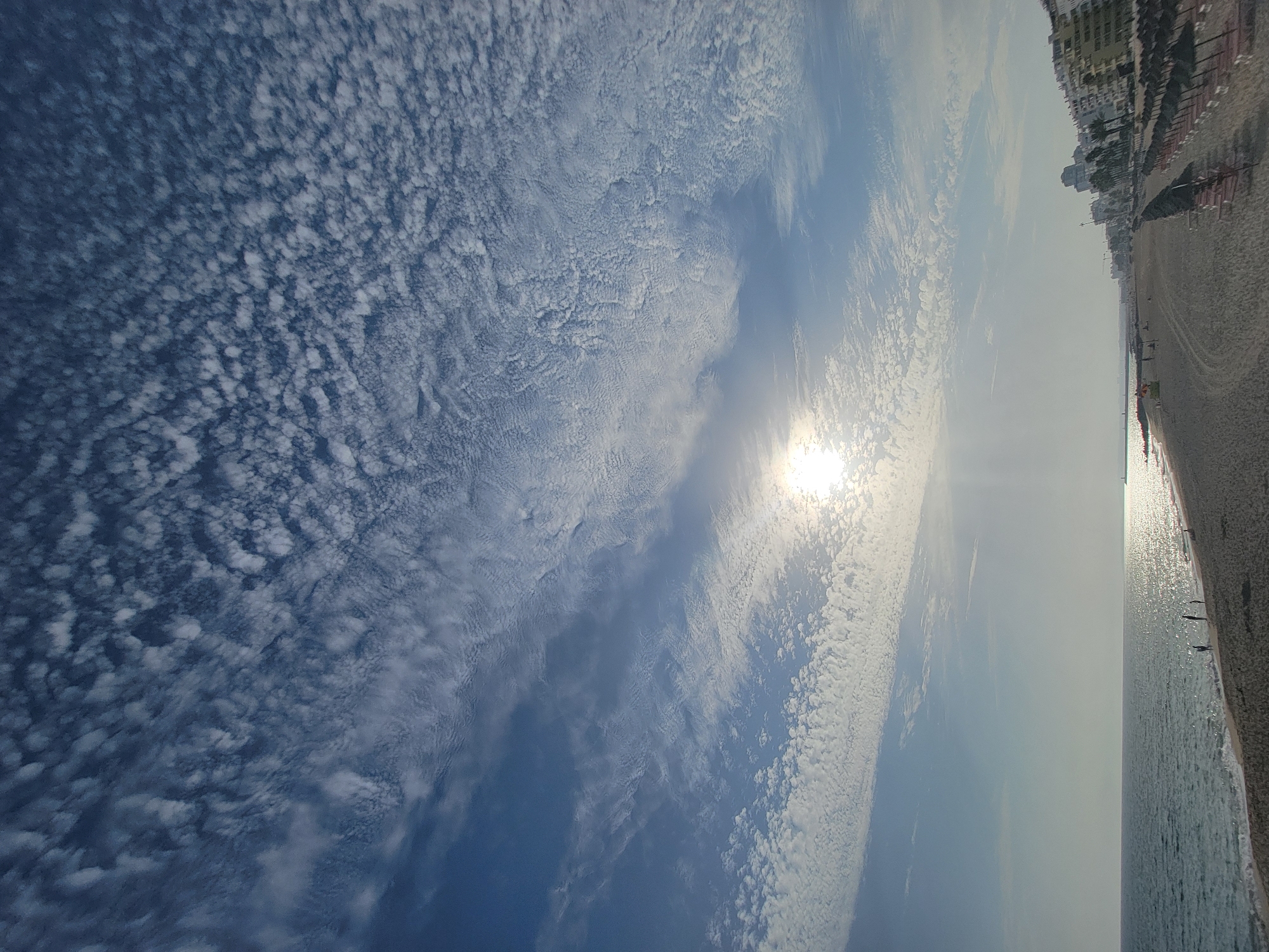 This media file depicts a beach scene in Quarteira, Portugal, oriented vertically, suggesting it was captured with the camera held sideways. The image features a vast expanse of blue sky filled with scattered, textured white clouds. The sun is prominently visible, partially obscured by clouds, casting a bright glow and creating a hazy effect. To the right, a sandy beach is visible, lined with rows of empty lounge chairs and umbrellas, indicating a daytime scene, likely outside of peak hours or off-season. Beyond the beach, there is a promenade with buildings and some palm trees, characteristic of a coastal resort town. The ocean is visible on the far right, with gentle waves lapping at the shore. A few small figures can be seen on the beach and in the water, suggesting minimal human activity. The overall mood is serene and bright, with a clear, sunny day. No visible text is present in the image.