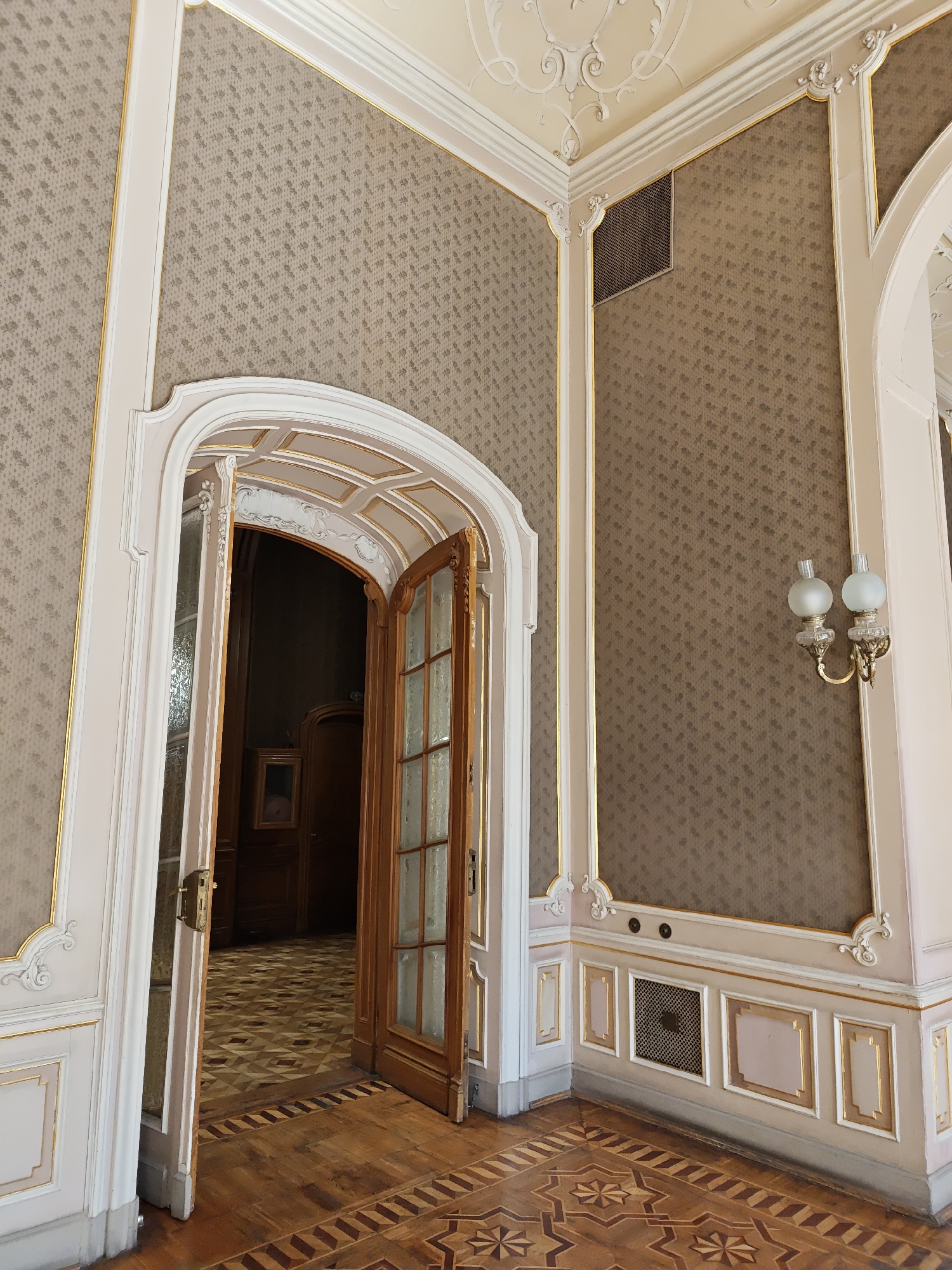 This image depicts the interior of an opulent room, likely in a historic building in Lviv, Ukraine. The focus is on a grand arched doorway, slightly ajar, revealing a glimpse into another room. The walls are adorned with intricate, repeating floral wallpaper in a muted brown hue, outlined by elegant white molding and accented with delicate gold trim. The ceiling features ornate plasterwork.
The doorway itself is a masterpiece of woodworking, with an arched top and wooden doors featuring glass panes. One door is partially open, showcasing a brass handle and intricate glass etching. Through the opening, a richly patterned parquet floor is visible, laid out in geometric designs of various wood tones. A dark wooden door is seen further into the adjacent room, along with a framed picture on the wall.
The room also features decorative elements such as a wall sconce with two frosted glass globes, an air vent in the upper wall, and decorative panels along the lower wall, also accented with gold trim. The floor in the foreground is also made of inlaid wood, displaying a complex star-like pattern. The overall impression is one of refined elegance and historical grandeur.