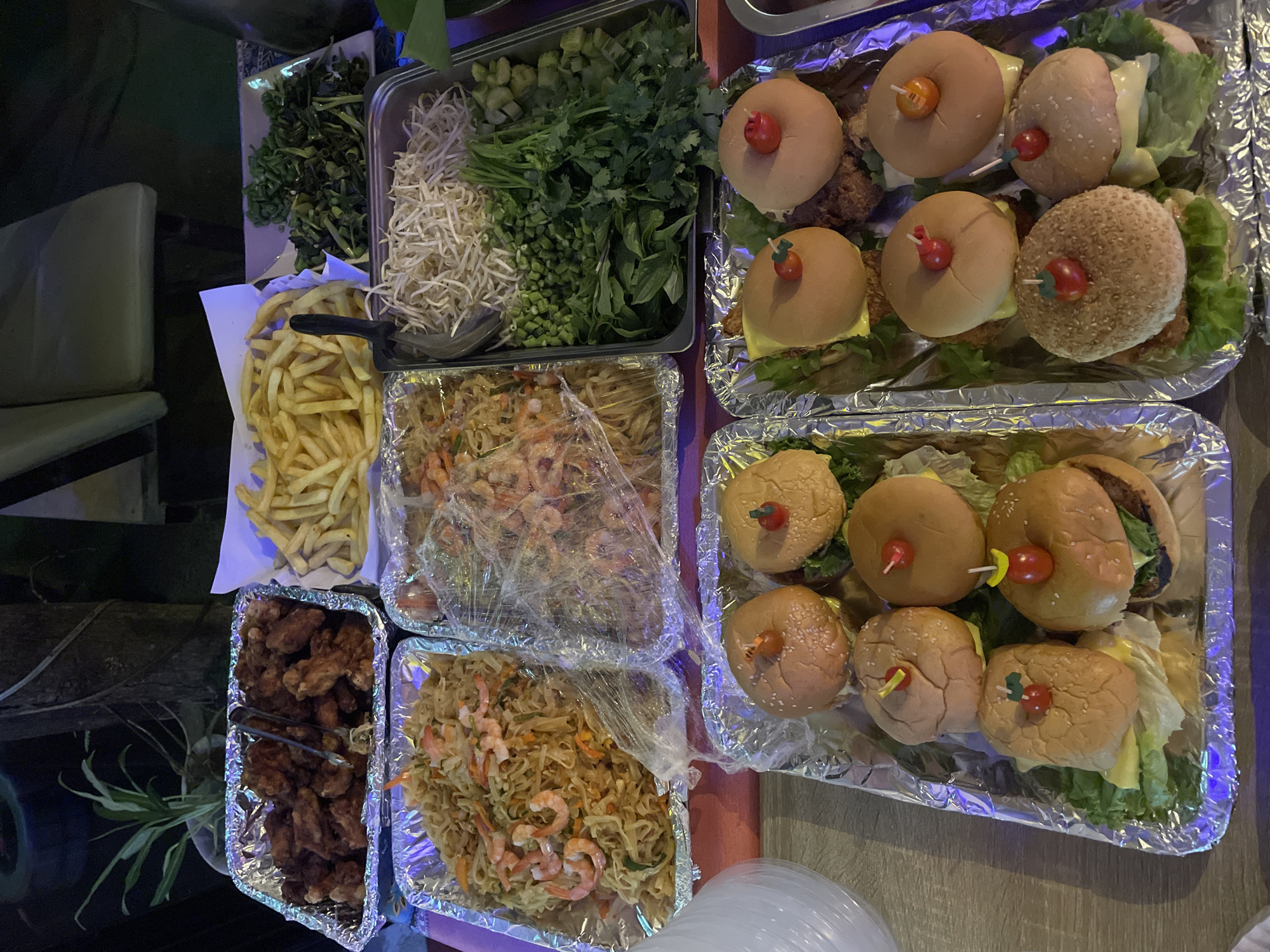 The image depicts a food spread at an outdoor event in Choeng Thale, Thailand. The food is arranged on aluminum foil-lined trays and platters, suggesting a casual buffet or catering setup. Visible food items include fried chicken wings, French fries, pad thai noodles with shrimp, bean sprouts, chopped green vegetables, and several mini burgers with cheese and lettuce, each topped with a cherry tomato. A serving utensil is placed in the bean sprouts. The background is dark, with hints of foliage, outdoor seating, and possibly artificial lighting, creating an evening ambiance. There are no people clearly visible in the frame.