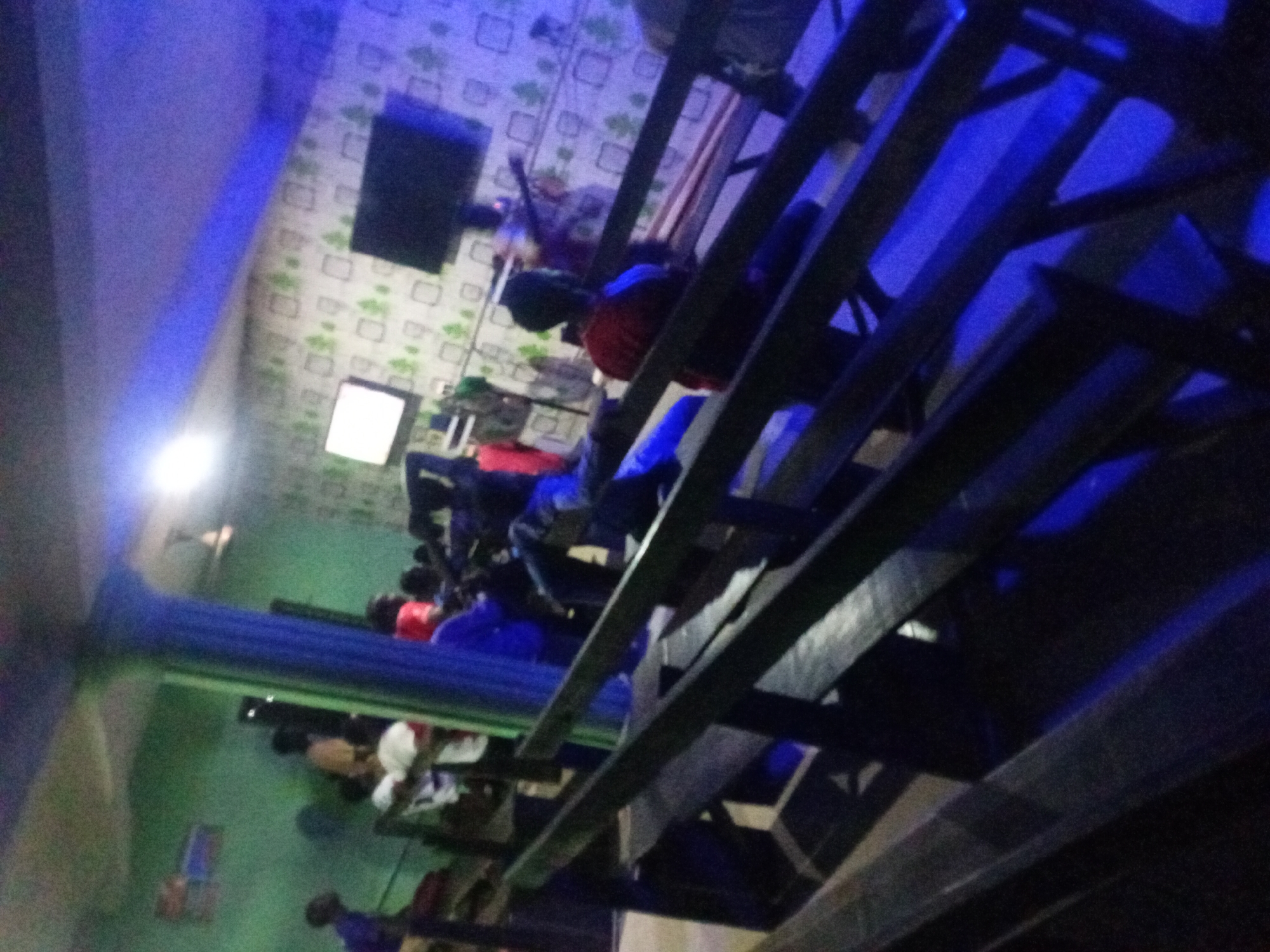 The image appears to be taken from an elevated position, possibly a balcony or upper level within a building. People are seated in rows, likely in a theater or presentation setting. There are monitors or screens on the wall, and blue lighting is prominent. The exact location and details about the people are unclear. The city is unknown.