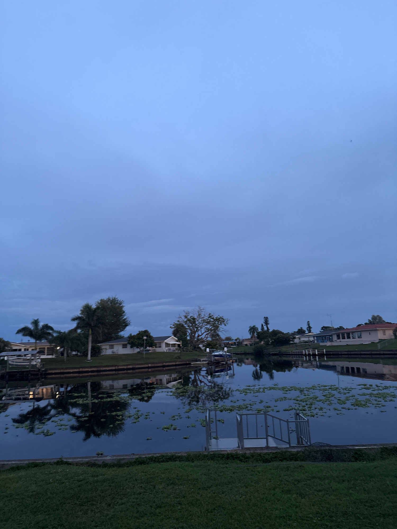 The image depicts a serene canal scene at dusk in North Port, Florida. The sky is a muted blue with scattered clouds, and the water reflects the surrounding houses and palm trees. A small metal dock with steps extends into the water, which is dotted with aquatic plants. Houses with white walls and tiled roofs line the canal, some with boats docked in front. The overall impression is one of tranquility and suburban living.