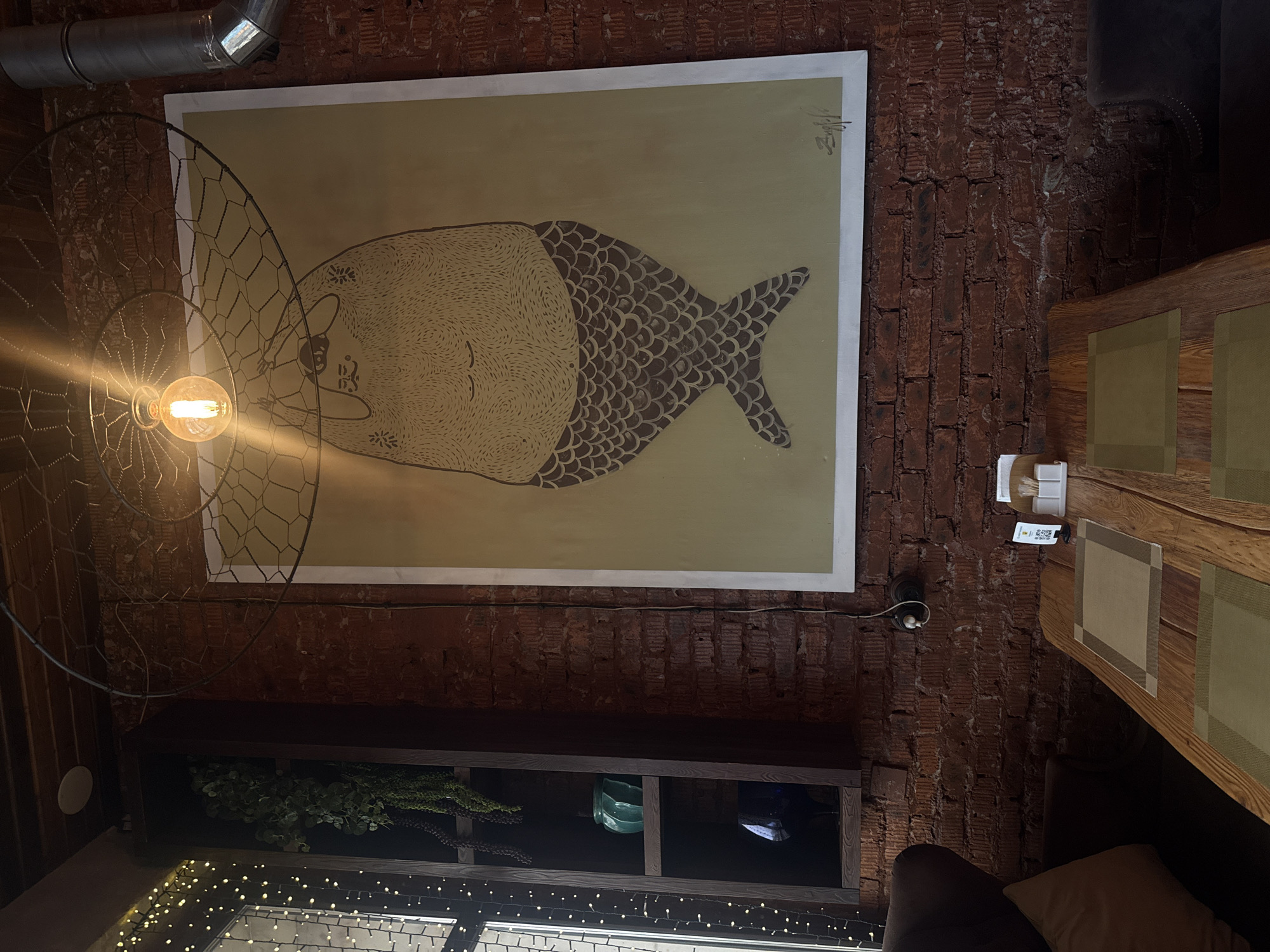 An interior shot, likely from a cafe or restaurant, showcases a rustic and cozy setting. The prominent feature is a large, framed artwork hung on an exposed red brick wall. The artwork depicts a stylized creature, resembling a fish with a human-like head, detailed with intricate line patterns and distinct scales on its tail. A signature, possibly "BophM" in cursive, is visible in the top right corner of the piece.
A large, open-cage light fixture with an exposed incandescent bulb hangs on the left, casting a warm, bright glow that partially illuminates the artwork. Above this, silver ventilation ductwork runs along the ceiling. To the right, a wooden table is set with two olive-green placemats. On the table, a white dispenser (possibly for napkins) and a small black tag with an unreadable QR code are present.
Below the artwork, a dark wooden shelf unit holds decorative items, including green plant-like elements and what appear to be glass bottles. Along the bottom edge of the image, a string of warm fairy lights adds to the inviting atmosphere. No people are visible in the scene, suggesting a quiet moment. The overall impression is one of warmth and comfort, typical of an evening setting in a casual establishment. The location context for this scene is Birobidzhan, Russia.