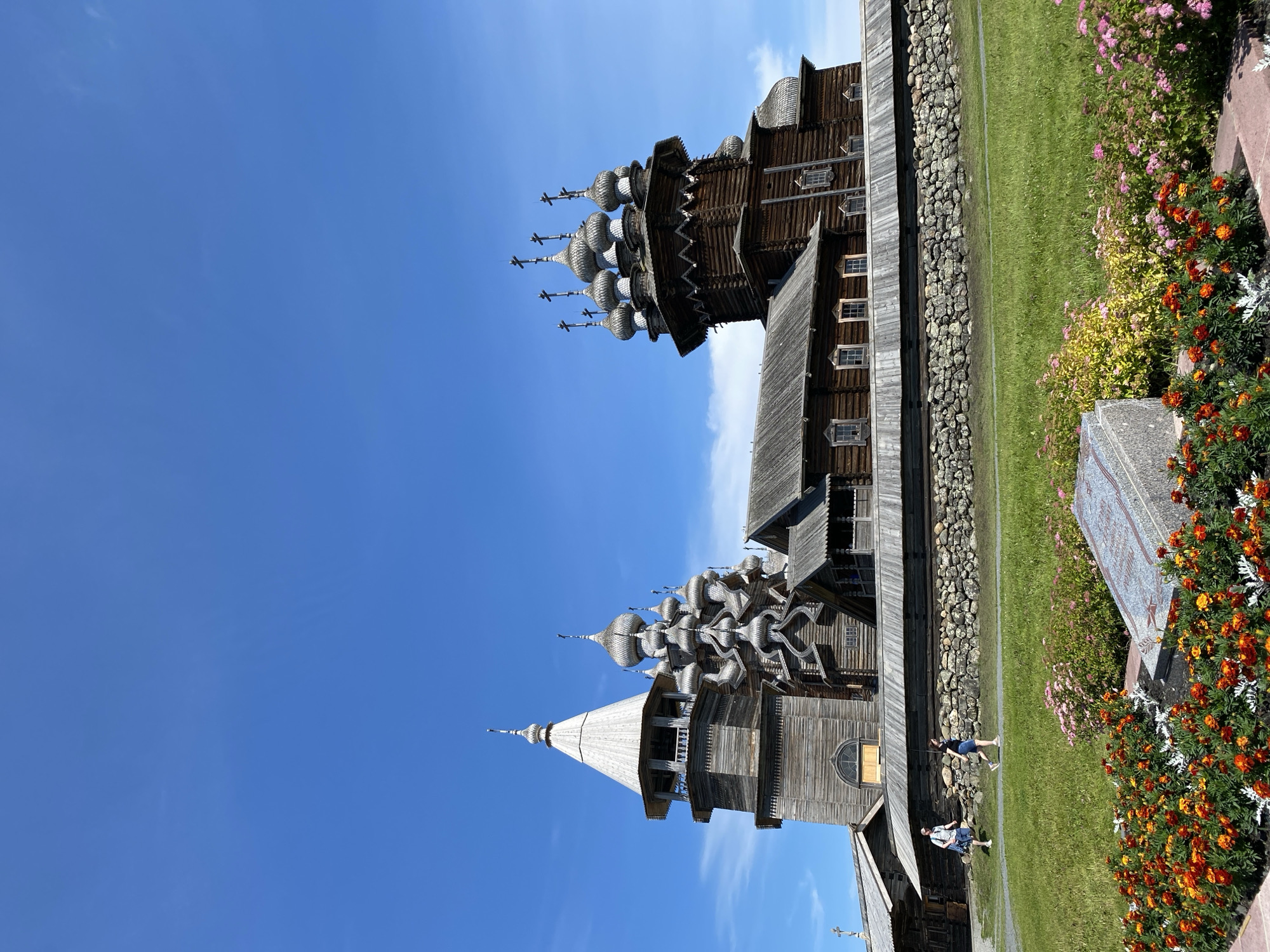 The image shows the Kizhi Pogost complex, a UNESCO World Heritage Site located on Kizhi Island in Karelia, Russia, which is near Boyarshchina. The complex features two wooden churches and a bell tower, all constructed in the traditional Russian architectural style. The larger church, the Church of the Transfiguration, is visible on the right with its distinctive onion domes, while the smaller Church of the Intercession and the bell tower are on the left. The buildings are made of wood and appear weathered, suggesting their age. They are surrounded by a low stone wall. In the foreground, there is a vibrant flower bed with orange and pink flowers, and a gravestone. Two individuals, a man and a woman, are walking on a path in the middle ground. The sky is a clear, bright blue with a few wispy clouds, and the sun appears to be shining, casting distinct shadows. The overall scene depicts an outdoor historical and cultural site under a pleasant weather.