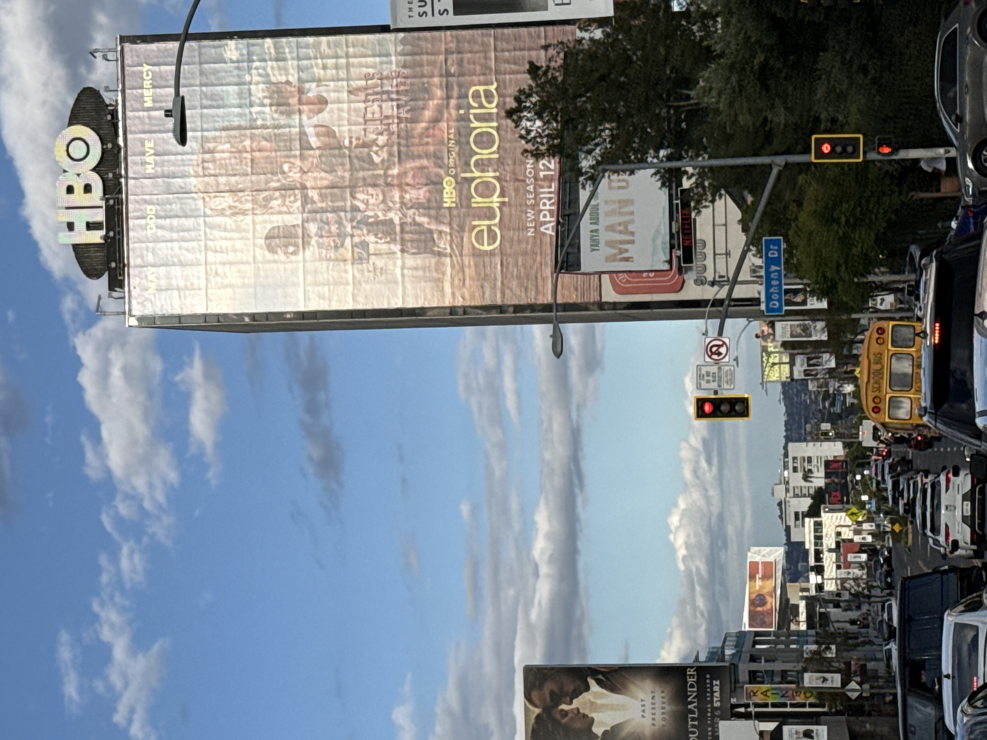 The image shows a street scene in West Hollywood, California. A large billboard displays an advertisement for HBO's "Euphoria," with the tagline "New Season April 12". Below that are additional billboards; one is an advertisement for "Man" starring Yahya Abdul-Mateen, and the other is an advertisement for "Outlander". A street sign indicates "Doheny Dr." Traffic is heavy with visible cars and a yellow school bus. The traffic light is red. A blue sky with scattered clouds forms the backdrop.