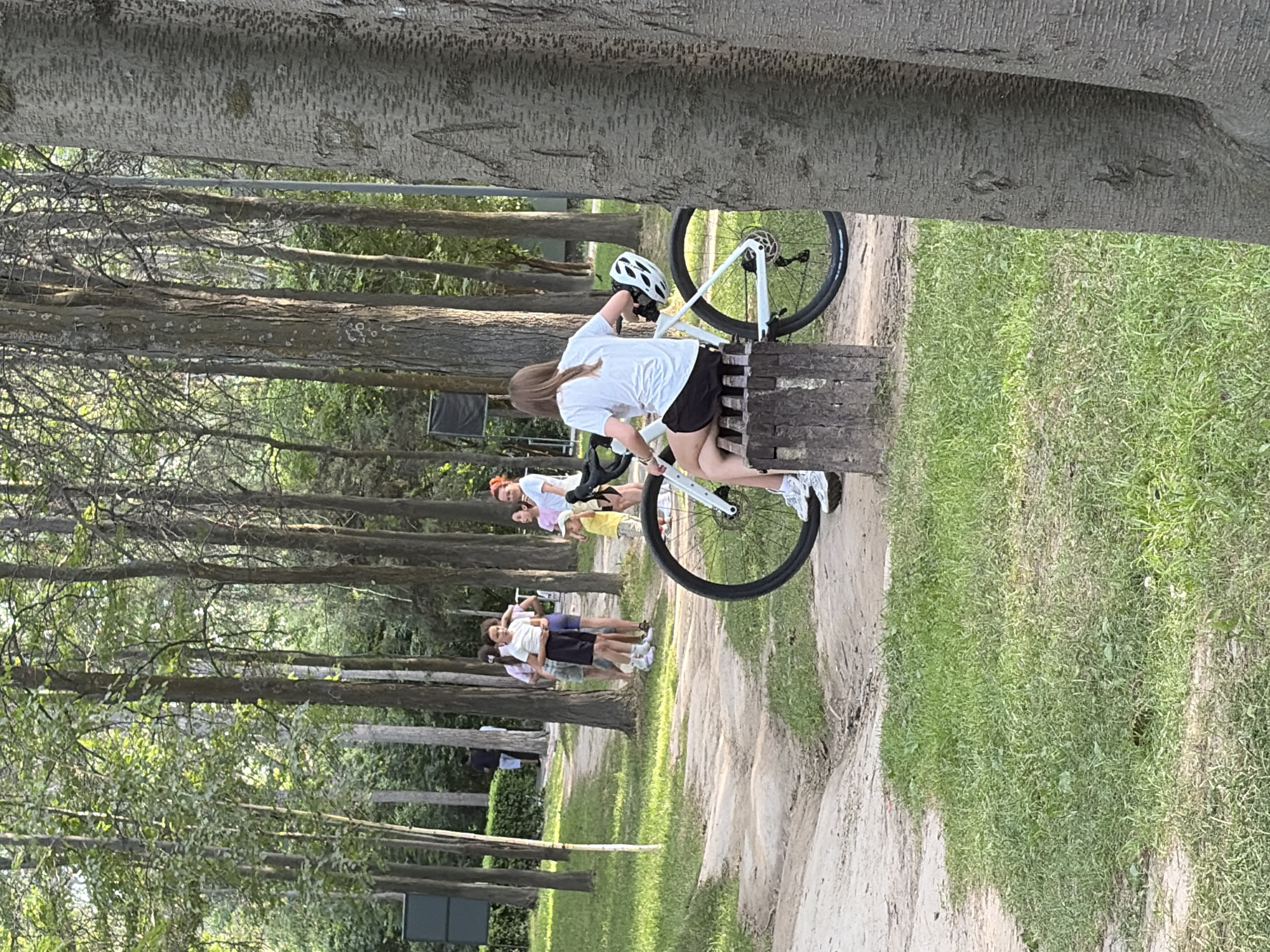 In an outdoor park setting with a gravel path winding through a wooded area, a young woman with blonde hair pulled into a ponytail is seated on a wooden bench next to a white bicycle. She is wearing a white t-shirt, black shorts, and white sneakers, and a white helmet rests on the bicycle's frame. She appears to be looking away from the camera, with her hands on the handlebars of the bicycle. In the background, a group of people, including several children and adults, are standing and conversing on the path. The environment consists of numerous tall trees with green foliage, and the ground is covered in grass and dirt. The overall scene suggests a casual gathering in a park.