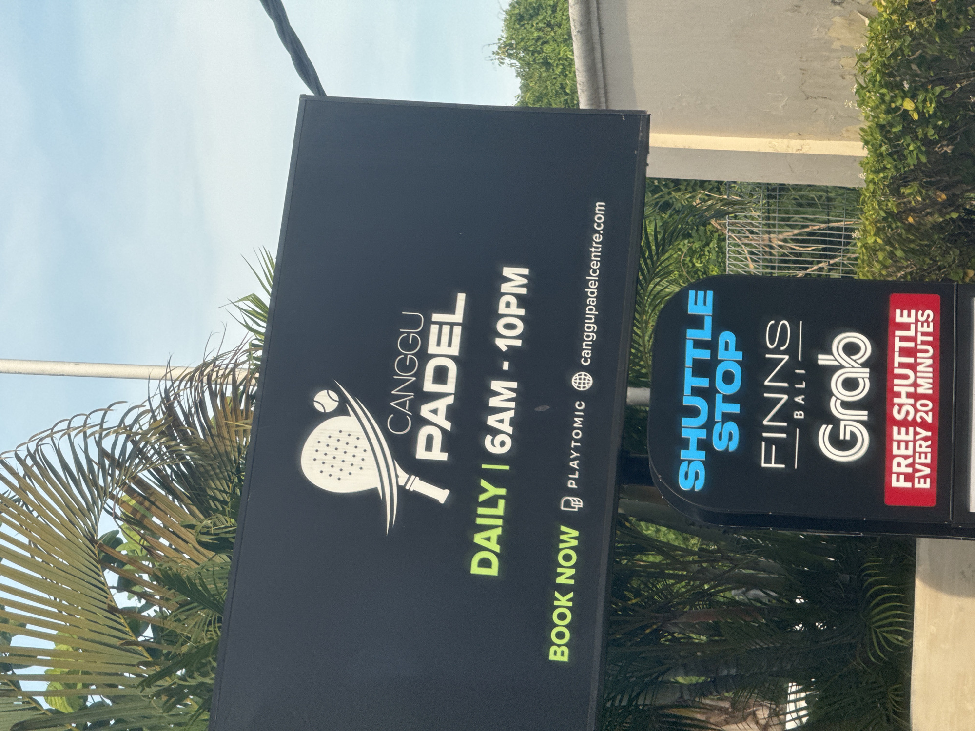 The image shows two advertising signs. The first sign on the left advertises "Canggu Padel" with operating hours "Daily | 6AM - 10PM." It also provides a booking option through Playtomic and the website canggupadelcentre.com. The second sign on the right indicates a "Shuttle Stop" for "Finns Bali" and "Grab," specifying "Free Shuttle Every 20 Minutes." These signs are presumably located in or near Canggu in North Kuta, Indonesia. There are trees and greenery in the background.