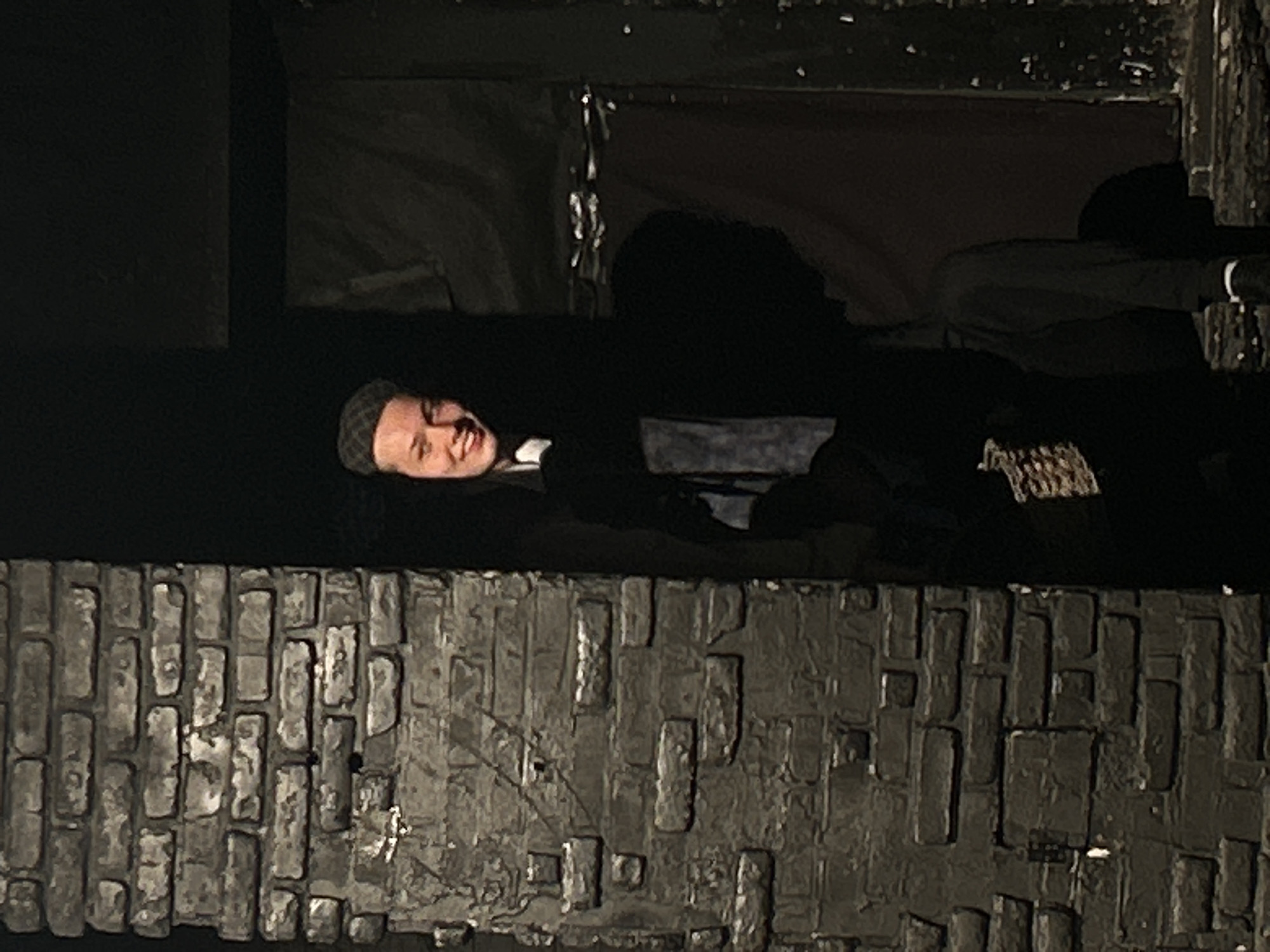 The image displays a theatrical or staged setting, centered on a dark doorway flanked by a brick-patterned wall.
**Location**: Unknown, Unknown.
**Environment**:
The left side of the frame is occupied by a vertical structure constructed to resemble a weathered dark grey brick wall. The bricks vary in shade and texture, showing some lighter highlights and visible mortar. To the right of this wall is a deep, dark opening or doorway. The background beyond this opening and to the right is largely obscured by darkness, though a dark, draped fabric is faintly visible in the upper right quadrant, and a reddish-brown element appears in the lower right, possibly part of another set piece.
**People**:
One individual is prominently featured, partially emerging from the dark doorway. This person, with fair skin, is seen from the chest up, smiling broadly towards the right side of the frame. They wear a dark, patterned flat cap, a white collar, and a dark outer garment, under which a blue or patterned tie or fabric is visible. Their expression is a wide, open-mouthed smile. Below and to the right of this person, a second, heavily shadowed figure is discernible as a dark silhouette. This obscured figure's head, shoulder, and a portion of their body are visible, appearing to be in a seated or hunched position, with minimal detail due to the low light.
**Actions**: The visible individual is leaning forward from the dark recess, directing their gaze and smile outward. The action of the obscured figure is not discernible beyond their posture.
**Lighting**: The scene is characterized by dramatic, low-key lighting. A primary light source illuminates the face and upper torso of the visible person, creating a stark contrast with the surrounding deep shadows. Portions of the brick wall are also dimly lit, highlighting their texture.