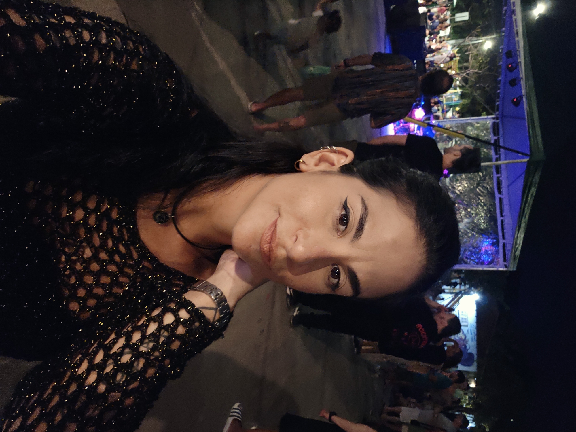 An eye-level selfie captures a woman in the foreground against the backdrop of a lively outdoor night event. The woman, with dark hair and a gentle smile, wears a black, shimmering, mesh-knit top and a necklace with a dark pendant. She has a metallic watch on her left wrist and multiple earrings on her right ear, including a helix cuff. Her gaze is directed towards the camera.
The background reveals a bustling scene at night, indicative of a public gathering, possibly a concert, festival, or market in Armação dos Búzios, Brazil. Numerous indistinct figures of people are visible, moving and congregating. To the right, a large blue tent or canopy structure is illuminated with bright blue stage lights. Other artificial lights dot the scene, suggesting a festive atmosphere. The ground appears to be a paved surface. One person in the background wears a shirt with the faint word "ROOTS" visible. The overall mood is casual and social.