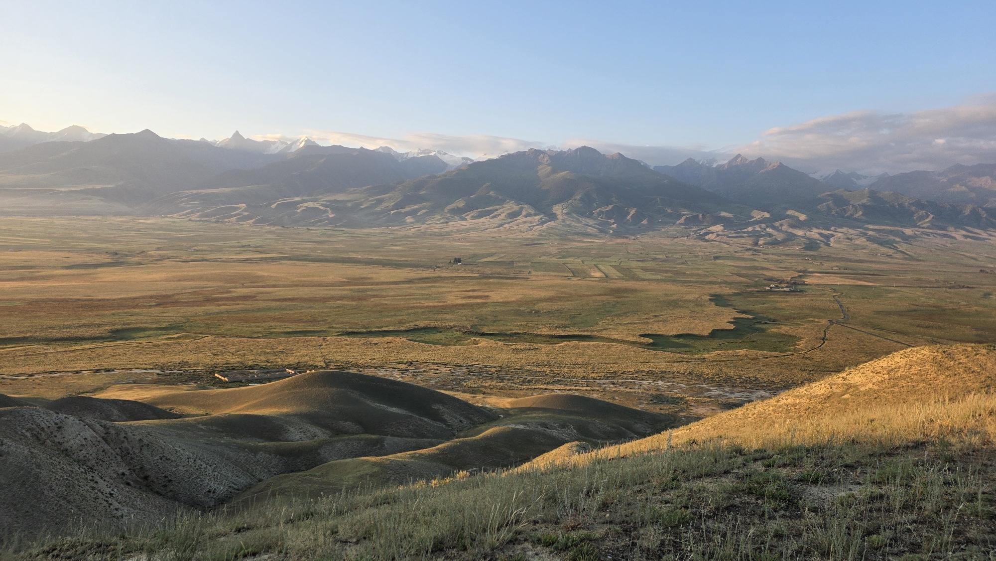 The image depicts a wide, panoramic view of a mountainous landscape under a clear, pale blue sky, possibly at dawn or dusk. The foreground shows a grassy hillside with dry, golden grass catching the warm light. Beyond the immediate foreground, rolling hills with undulating contours, cast in shadow and bathed in soft light, dominate the mid-ground. These hills transition into a vast, flat valley that stretches towards a range of imposing mountains in the background.
The valley floor is a patchwork of light and dark green and golden-brown hues, suggesting a mix of vegetation and perhaps agricultural fields. A winding river or stream is visible in the valley, carving a dark, serpentine path through the landscape. Scattered in the distance within the valley, there appear to be small clusters of buildings, indicating human settlement. The mountains in the background are rugged and majestic, with snow-capped peaks glinting in the sunlight, indicating high altitude. The overall impression is one of natural beauty, vastness, and tranquility.
The prompt mentions the location as Ак Жар, Kyrgyzstan. While the image itself does not contain explicit markers of this specific location, the terrain and mountain range are consistent with the geography of Kyrgyzstan, a country known for its mountainous landscapes and valleys. There are no people, animals, or specific objects that would suggest a particular event or narrative beyond the natural environment. The scene is serene and appears untouched by significant human activity, except for the distant structures in the valley.