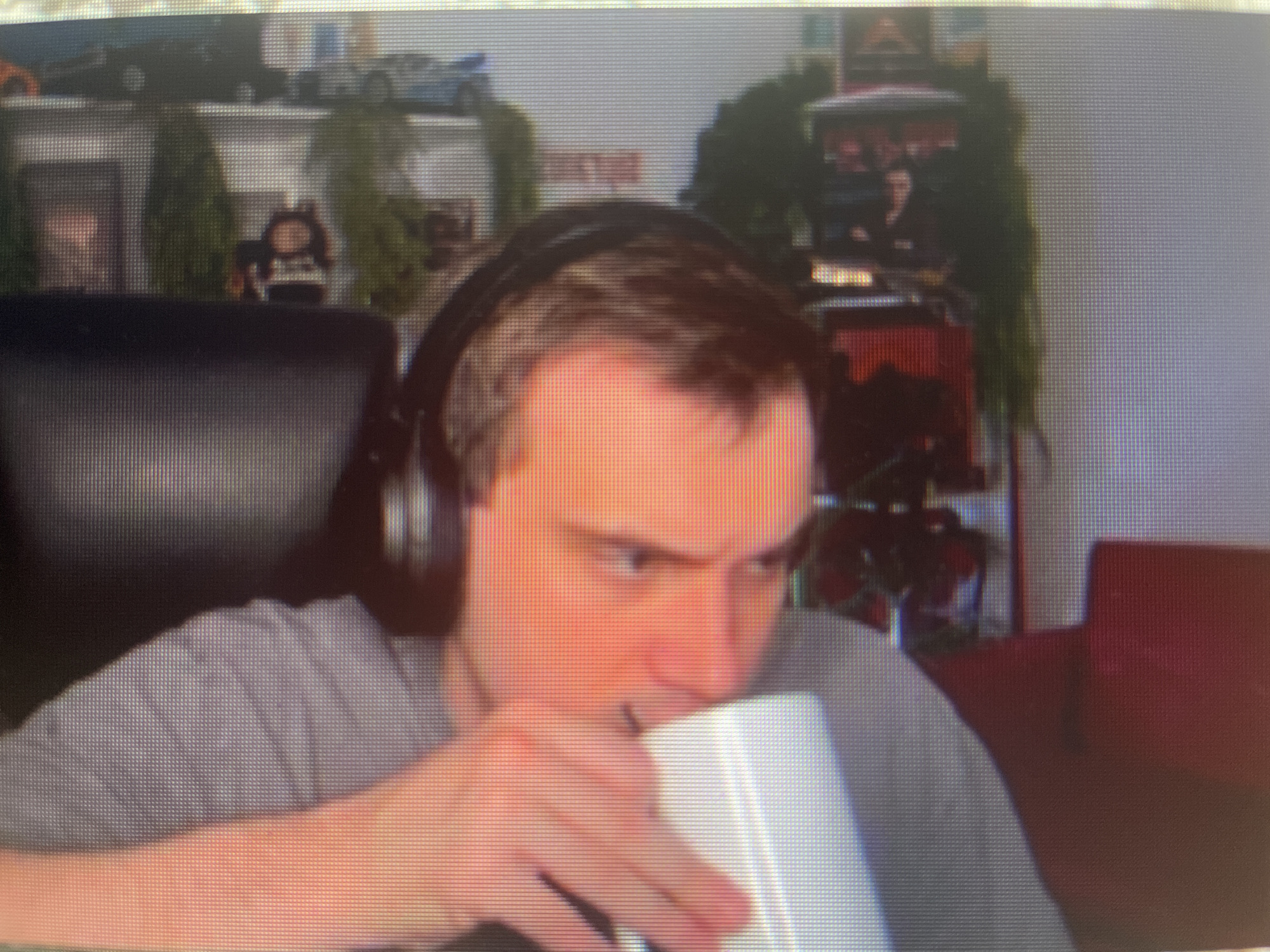 A man wearing headphones is sitting in a chair, holding a white mug and drinking from it. He is wearing a gray t-shirt. In the background, there are shelves with various items, including toy cars, plants, and figurines. A red chair is visible to the right of the man. The image appears to be a screen capture from a video or stream. The location is not identifiable from the image.