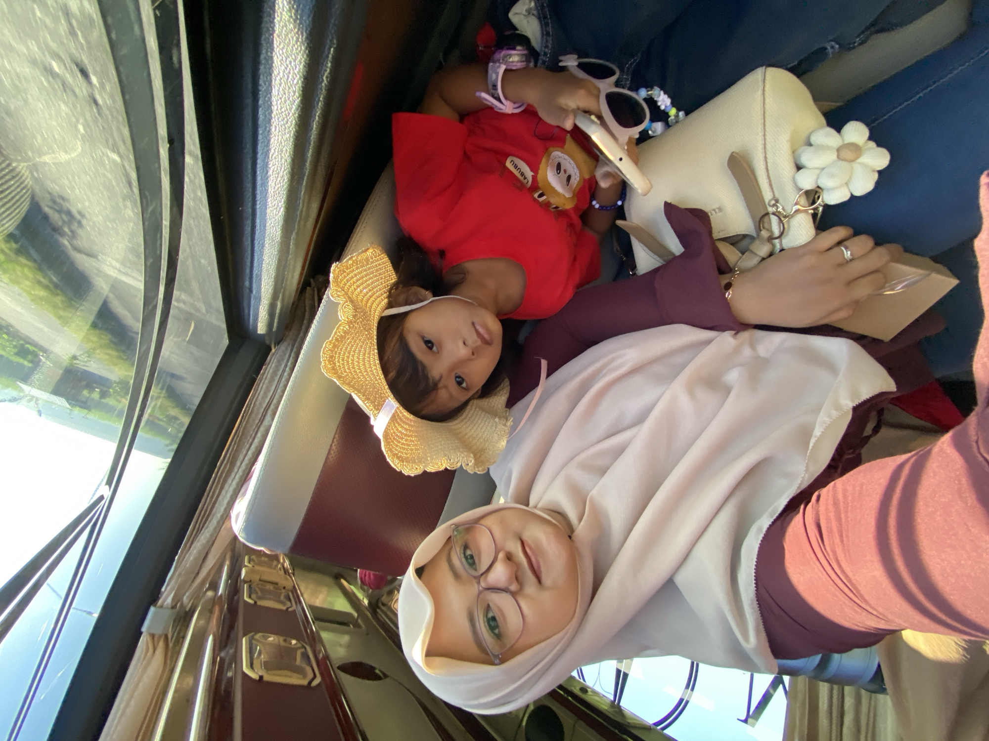Inside a vehicle, an adult female and a young child are observed in Pamotan, Indonesia. The adult female, on the left, wears a light beige hijab, glasses, and a maroon top, subtly smiling at the camera, indicative of a selfie. Her left hand, wearing a ring, rests on her lap near a beige handbag with a white floral charm, holding a small brown object. To her right, a young child wears a straw hat, a red t-shirt with a "LABUBU" character, and dark jeans, holding a white smartphone horizontally. The child has a neutral expression, and pink-framed sunglasses are visible nearby. The vehicle interior features red and white seats, overhead compartments, and beige curtains tied back from the windows. The exterior view through the windows reveals a bright, sunny day with a road, green vegetation, and distant hills or structures.
