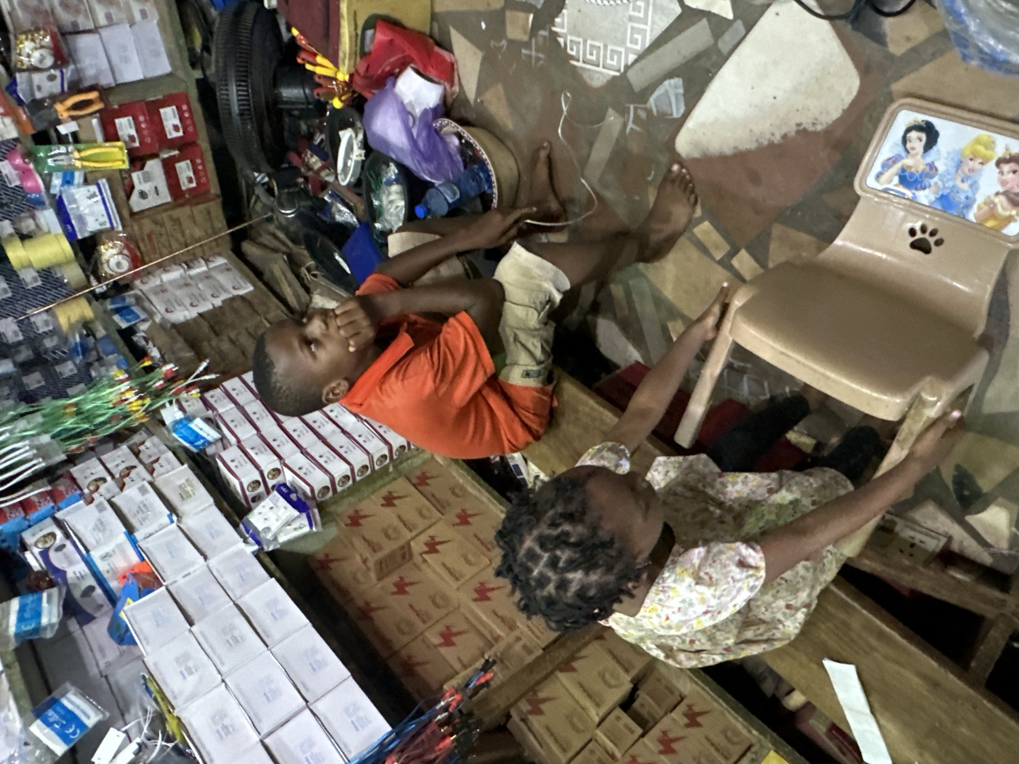 In a shop in Narayi, Nigeria, two young children are seen. A boy, appearing to be around seven years old, sits on a wooden bench, leaning his chin on his hand and looking to the left. He wears an orange polo shirt and beige shorts. Beside him, a younger girl with braided hair sits on the same bench. She is wearing a floral-print dress and is reaching towards a light brown plastic chair. The shop appears cluttered, with shelves in the background stocked with various items, including boxes, electrical components, and tools. The floor is tiled with a patterned design. A plastic bottle of water and other miscellaneous items are visible near the children.