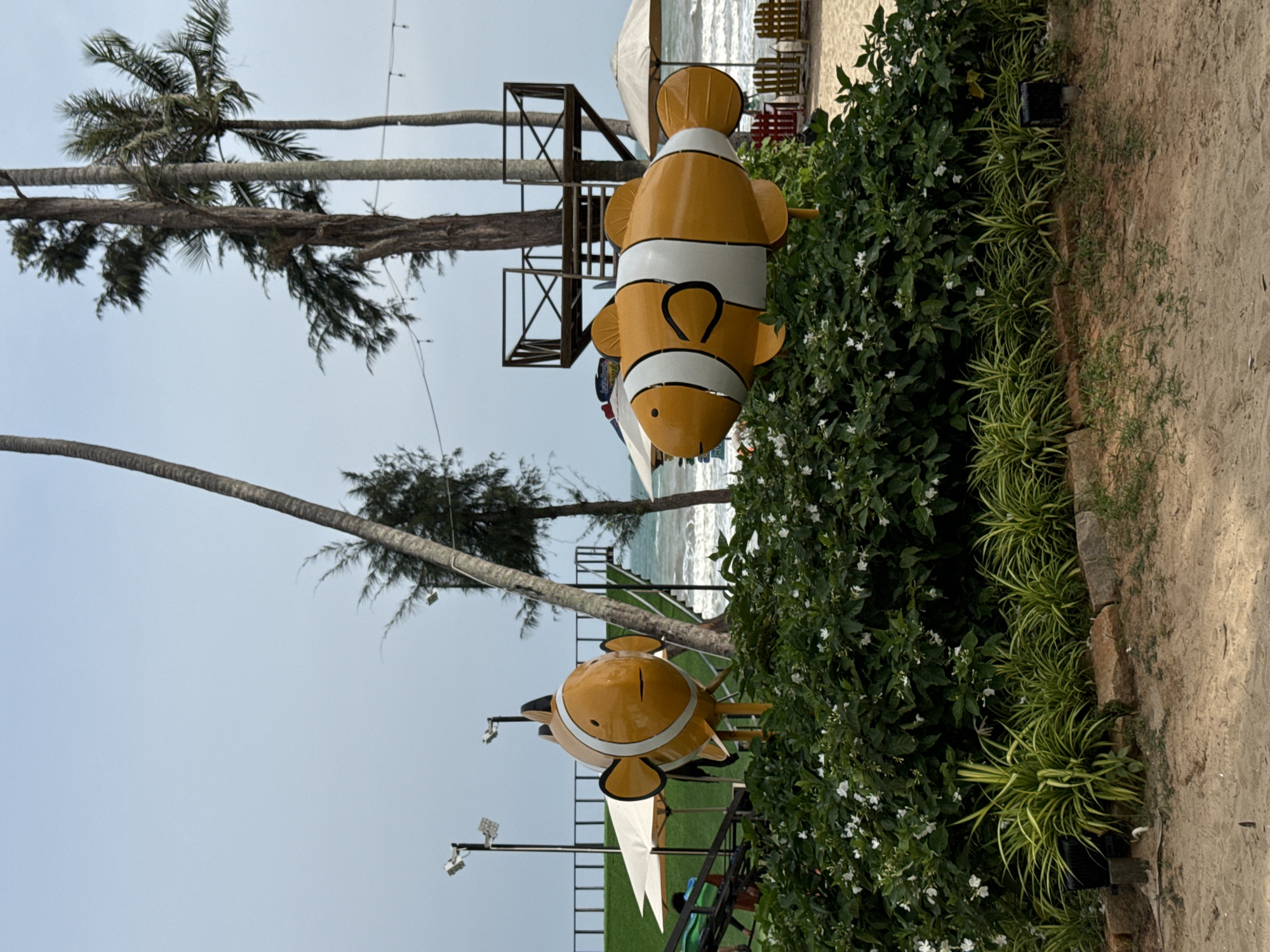 The image depicts an outdoor scene with two large, ornamental clownfish statues in the foreground, positioned amidst lush green bushes with white flowers. Behind the clownfish, a beach environment is visible with palm trees, a wooden structure resembling a treehouse, and umbrellas. The ocean is in the background with waves. The scene is likely in Mũi Né, Vietnam, a coastal resort town known for its beaches and tourist attractions. The overall atmosphere is tropical and relaxed.