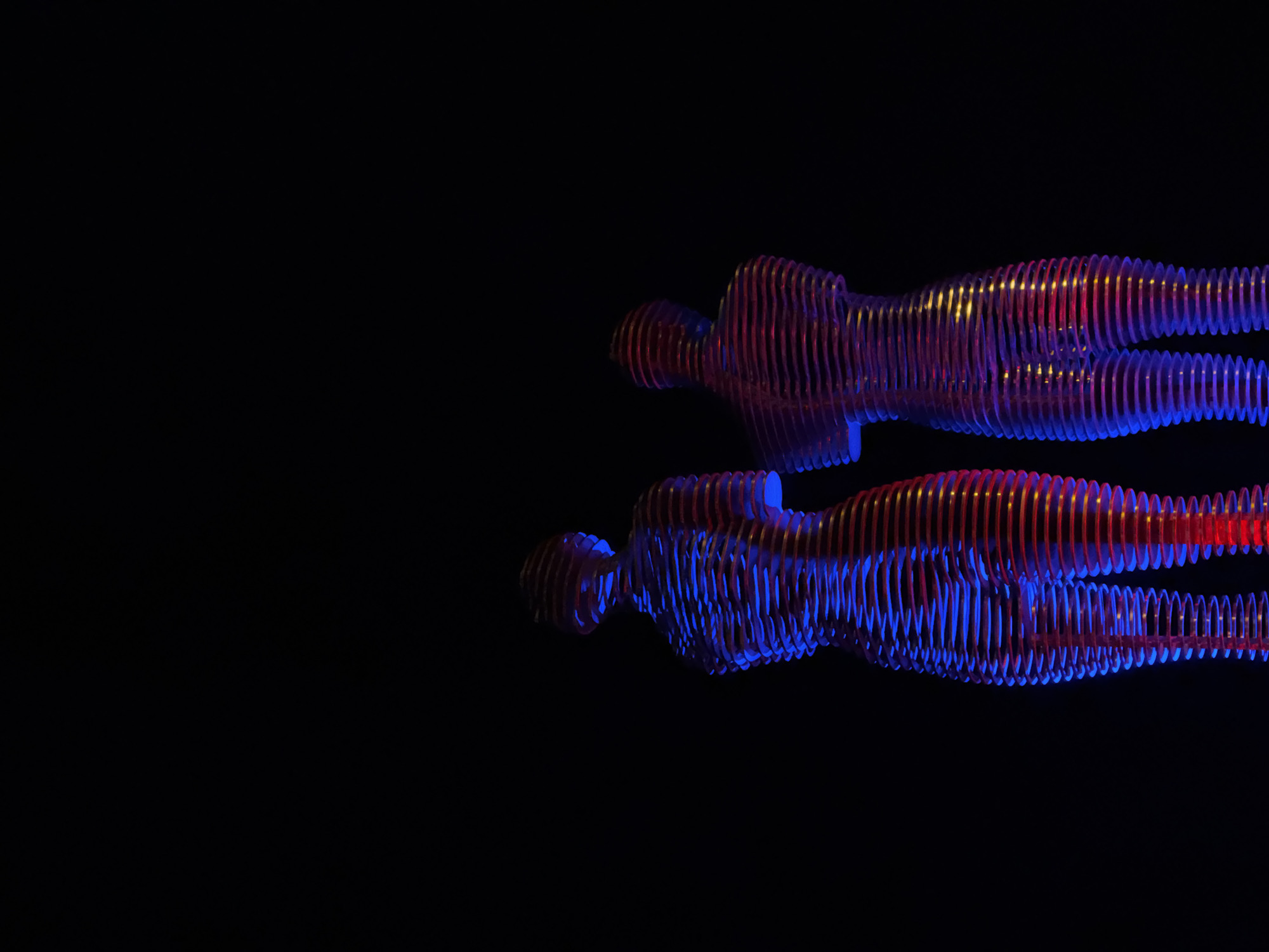 Two abstract, illuminated sculptures of human figures stand against a black background. The sculptures are made of horizontal, stacked rings that create a layered effect. They are lit with alternating bands of red and blue light, which create a translucent appearance and cast shadows. The figures are positioned side-by-side, with one slightly behind the other, and appear to be facing away from the viewer. These sculptures are known as "Ali and Nino," a famous monument located on the coast of Batumi, Georgia. The monument represents a love story between a Muslim boy from Azerbaijan and a Georgian princess.