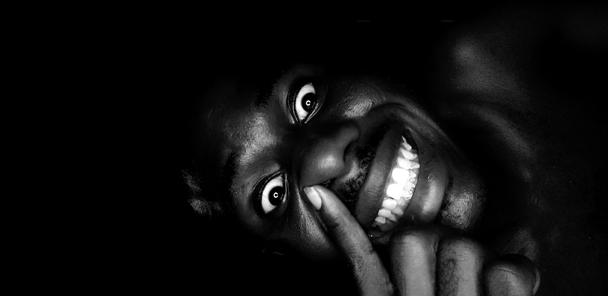 A black and white close-up photograph depicts a dark-skinned person with wide eyes, bared teeth, and a finger touching their upper lip. The subject's head is tilted, and the background is primarily dark and indistinct. There is no information to support the location being Narayi, Nigeria.