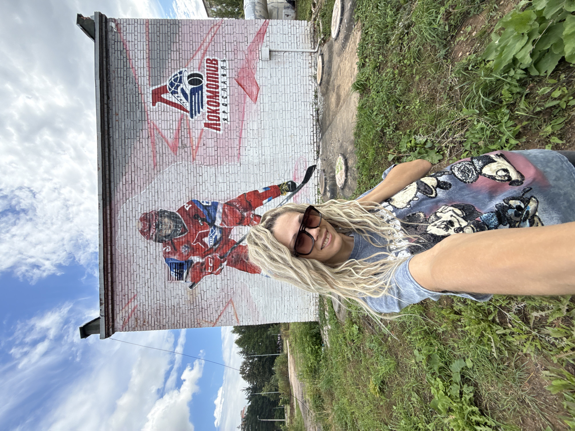 A woman with blonde dreadlocks, wearing a blue t-shirt with a graphic print and large sunglasses, takes a selfie in front of a brick building with a mural. The mural depicts a hockey player in a red and blue uniform and the logo for "LOKOMOTIV YAROSLAVL". The building is located in Rusanovo, Russia, and is surrounded by green grass and trees. The sky is blue with some clouds. There are several manhole covers visible on the ground in front of the building.