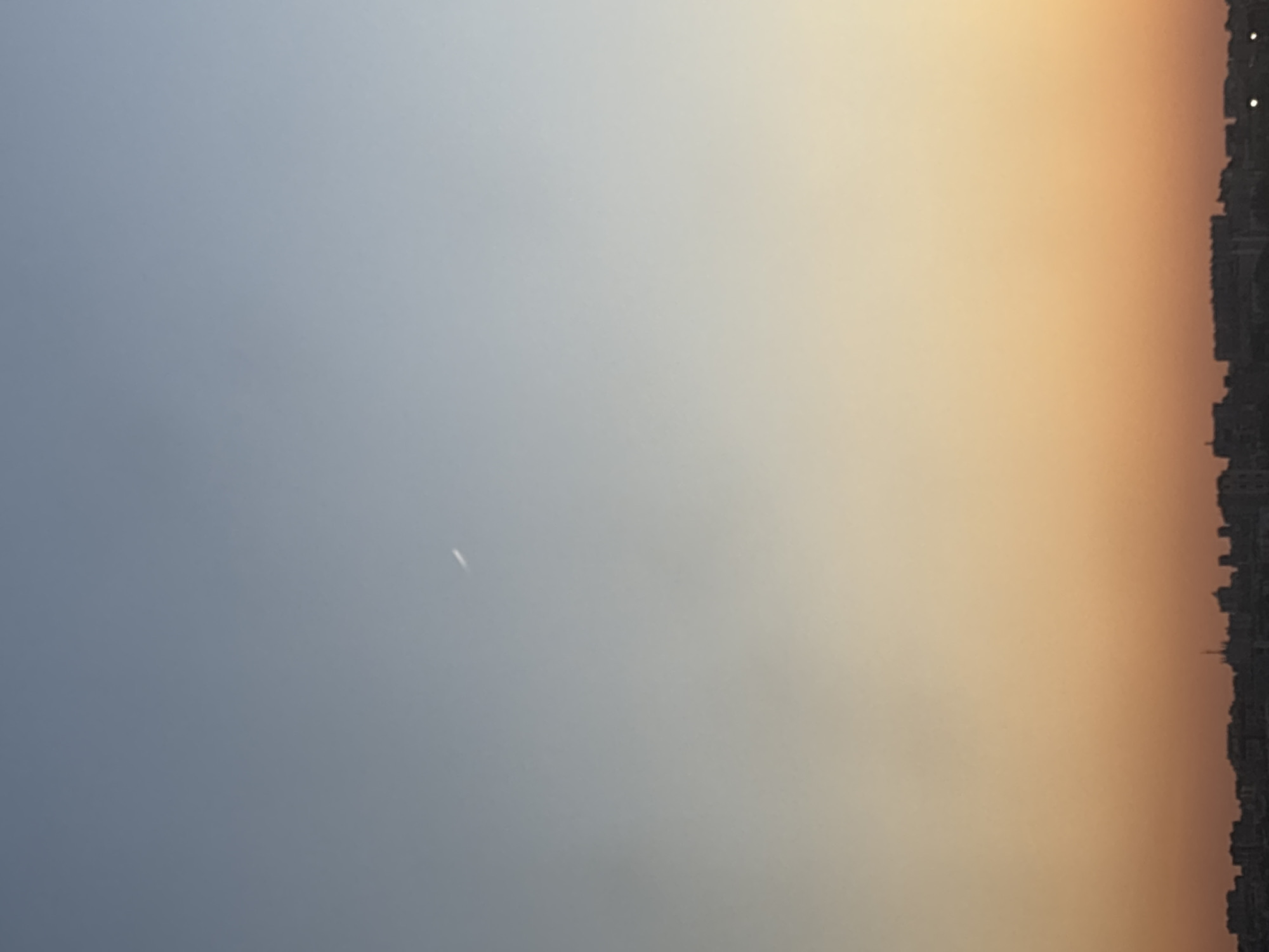 The image shows a cityscape against a gradient sky, transitioning from a blue-grey hue on the left to orange on the right, suggesting sunset or sunrise. A thin, bright streak is visible in the sky, potentially a plane or contrail. The city silhouette on the right comprises buildings of varying heights and architectural styles. Without further information, identifying the exact location within Vsevolozhsk, Russia is not possible.