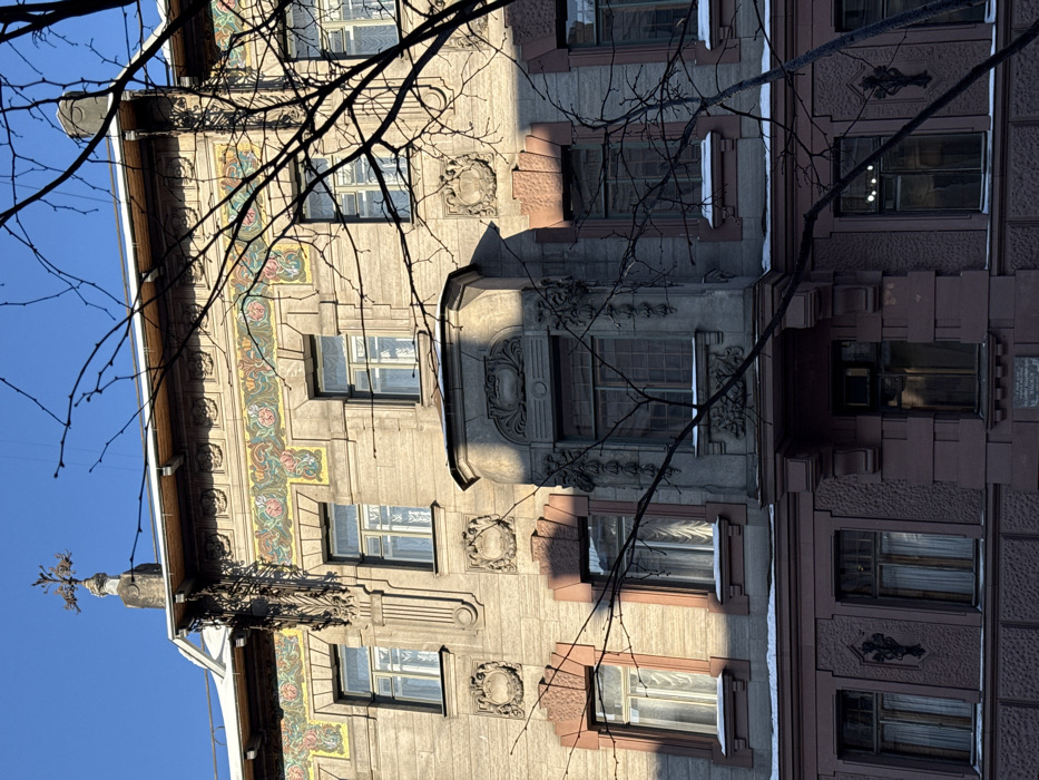 This image displays a detailed, partial view of the facade of the House of Vasilyeva-Khomutova, located at 72 Bolshoy Prospekt P.S. (Bolshoy Prospekt Petrogradskoy Storony) in Saint Petersburg, Russia.
The building is an example of early 20th-century Art Nouveau (Modern style) architecture. The visible section spans approximately three to four stories. The upper floors are constructed of a light grey/beige stone or stucco, while the lowest visible floor features a darker reddish-brown, rusticated finish.
A prominent, colorful floral frieze, featuring stylized pink roses, green foliage, and yellow accents, runs horizontally between the upper two main floors. Below this frieze, the facade includes multiple rectangular windows with white frames, many of which have small accumulations of snow on their sills. Some windows reveal sheer white curtains behind the glass.
Central to the composition is an ornate, grey stone bay window on the second floor (from the bottom up), elaborately decorated with sculpted floral and foliate motifs. Above the roofline, a distinctive, dark-colored attic ornament resembling a stylized metal tree or plant rises from a stone base. A vertical, decorative relief element is positioned to its left.
The lower reddish-brown section of the facade features additional windows and a rectangular plaque, partially obscured by shadows, on the left side of the second bay from the left.
The scene is illuminated by strong sunlight from the right, casting distinct shadows on architectural elements and highlighting textures. The sky is clear blue. Bare, dark tree branches partially obstruct the view of the right side of the facade. Snow is visible on horizontal ledges, window sills, and portions of the roof, indicating a winter setting. No individuals, actions, or interactions are visible.