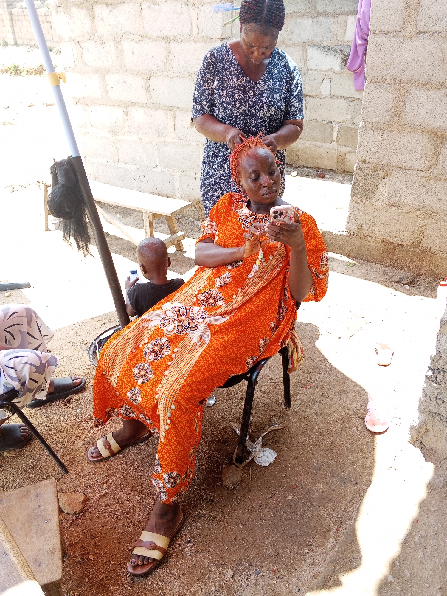 The image captures an outdoor scene in an unidentified city. The central subject is a woman with orange-tinted hair, seated on a black chair, engrossed in her smartphone. She is clad in an orange patterned dress and brown sandals. Another woman, standing behind her, is braiding her hair. A small child is visible behind the woman in the orange dress, holding a white bottle.
To the left of the scene, a black hairpiece hangs from a pole. The backdrop is a rough, light-colored brick wall. A small wooden bench sits against the wall. The ground is dirt. Additional objects include a pink slipper, trash, and furniture legs.
