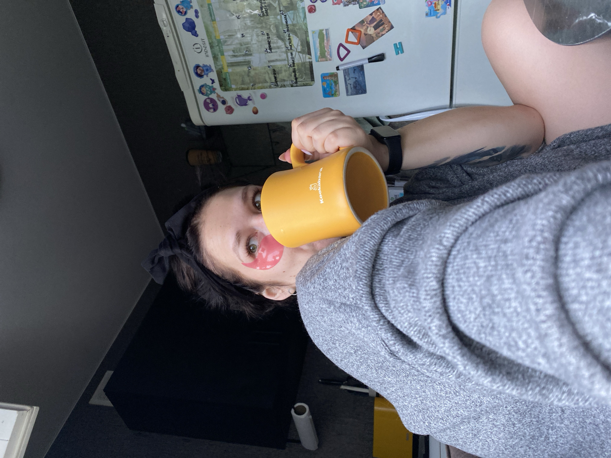 A woman is indoors, wearing a grey sweater and drinking from a yellow mug, with a pink eye mask under her right eye. The mug has the words "Kookaburra" printed on it. She has a black hair tie in her hair and a black smartwatch on her left wrist. There is a tattoo visible on her arm. Behind her is a white refrigerator covered with magnets, including a calendar. In the background, there is a dark-colored surface, possibly a countertop or cabinet, and a grey wall. A roll of paper towels lies on the floor. Based on the presence of the "Indesit" logo on the refrigerator and the "Kookaburra" mug, it's possible that the location is in a residential area where these products are available. Given the prompt mentioning Chaikovsky, Russia, it's reasonable to assume this image was taken there.