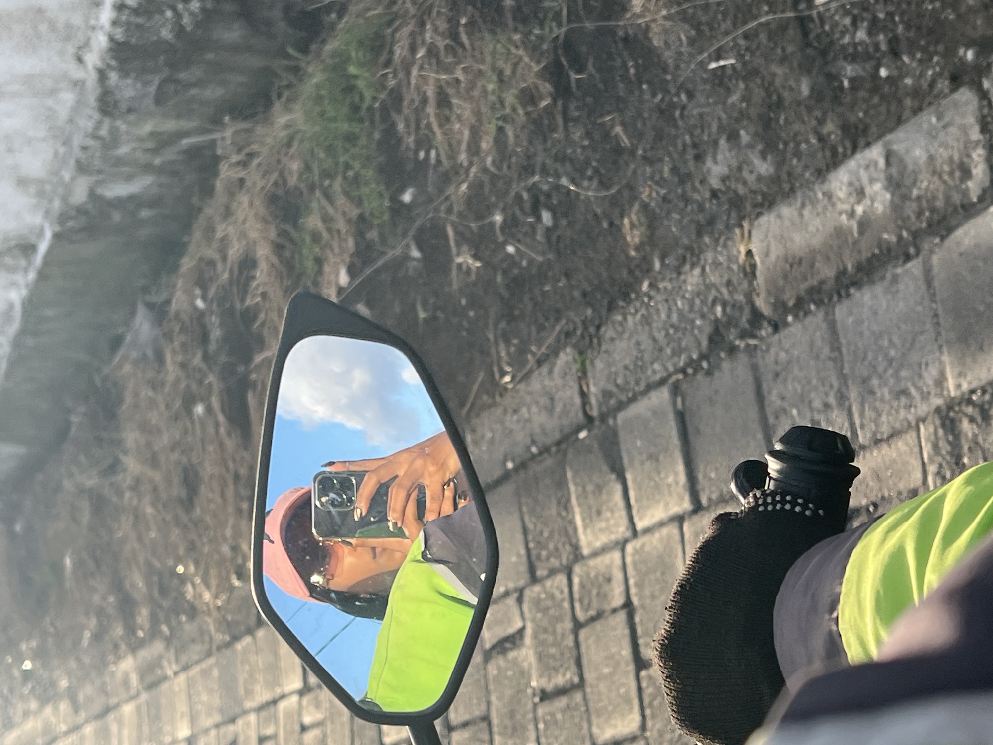 A person is taking a selfie in the mirror of a motorcycle. The person is wearing a pink cap, sunglasses, and a bright green vest. They are holding a smartphone in their right hand. The background shows a paved walkway and dry vegetation. The scene appears to be outdoors, possibly in a city environment. The location is identified as North Kuta, Indonesia.