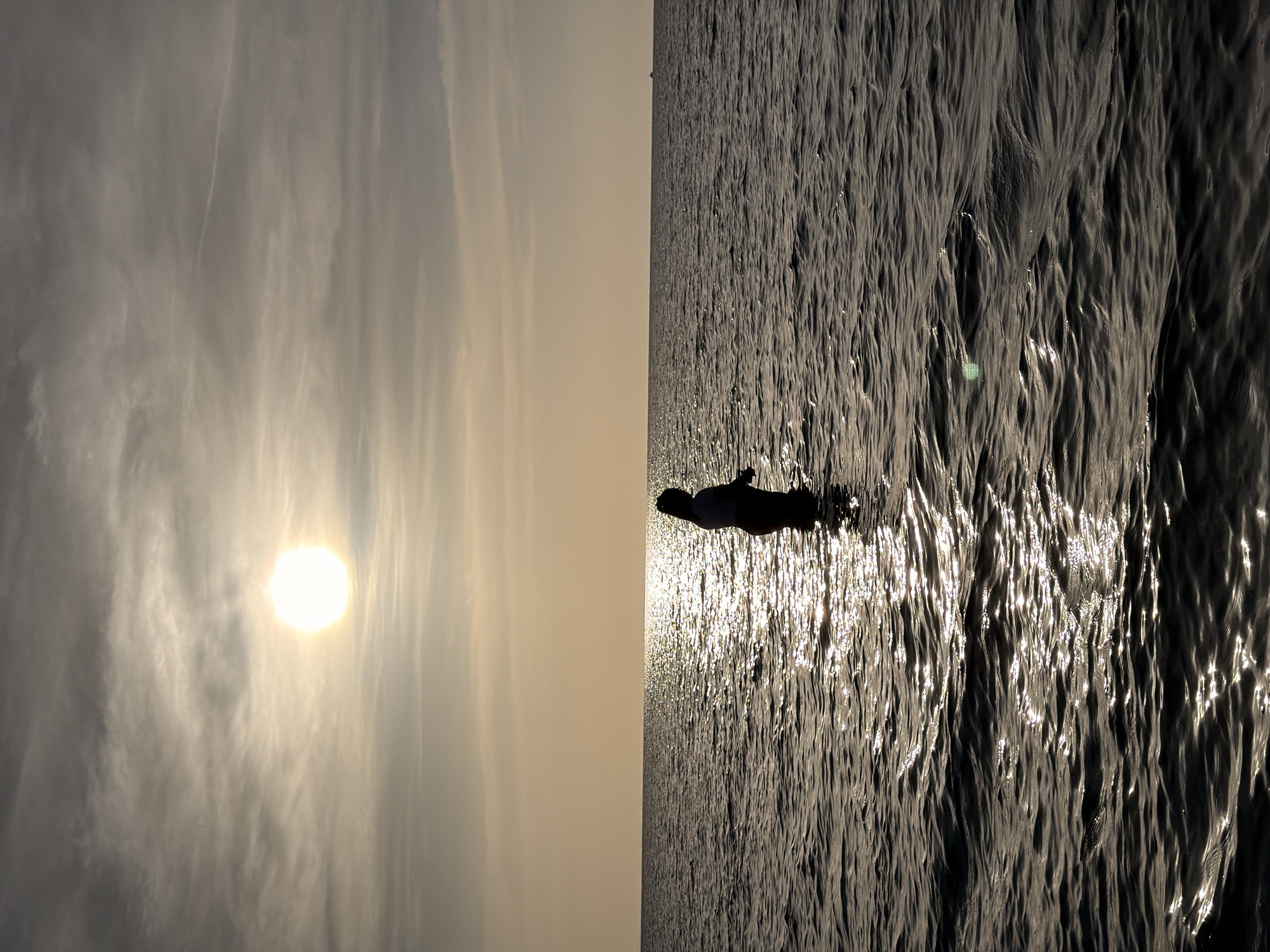 A silhouette of a person stands in the ocean with the sun setting in the background, creating a shimmering reflection on the water. The person is facing away from the camera, and their outline is clearly defined against the bright sky. The sky is filled with wispy clouds, and the sun, a bright orb, is positioned in the upper left quadrant of the image. The ocean appears calm with gentle ripples. This scene likely depicts a sunset at a beach in Ko Samui District, Thailand.