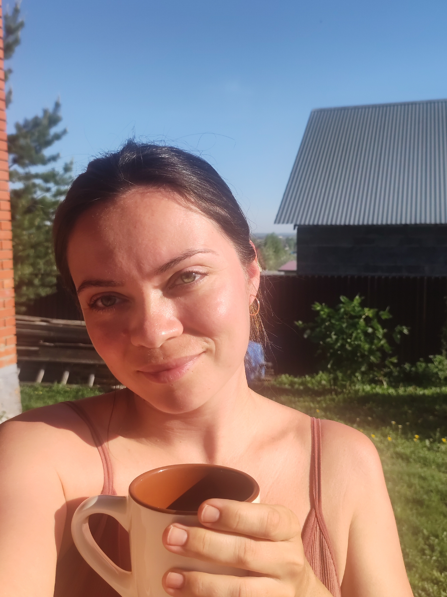 A young woman is holding a mug in her right hand, likely outdoors. The mug has a cream-colored exterior and a brown interior. The woman has dark hair pulled back and is wearing a sleeveless top. She is looking at the camera with a slight smile. Behind her is a brick wall on the left and a building with corrugated metal roofing on the right. The background includes trees and green grass. The setting appears to be near Фоки, Russia, based on the style of architecture. The time of day appears to be daytime with bright sunlight.