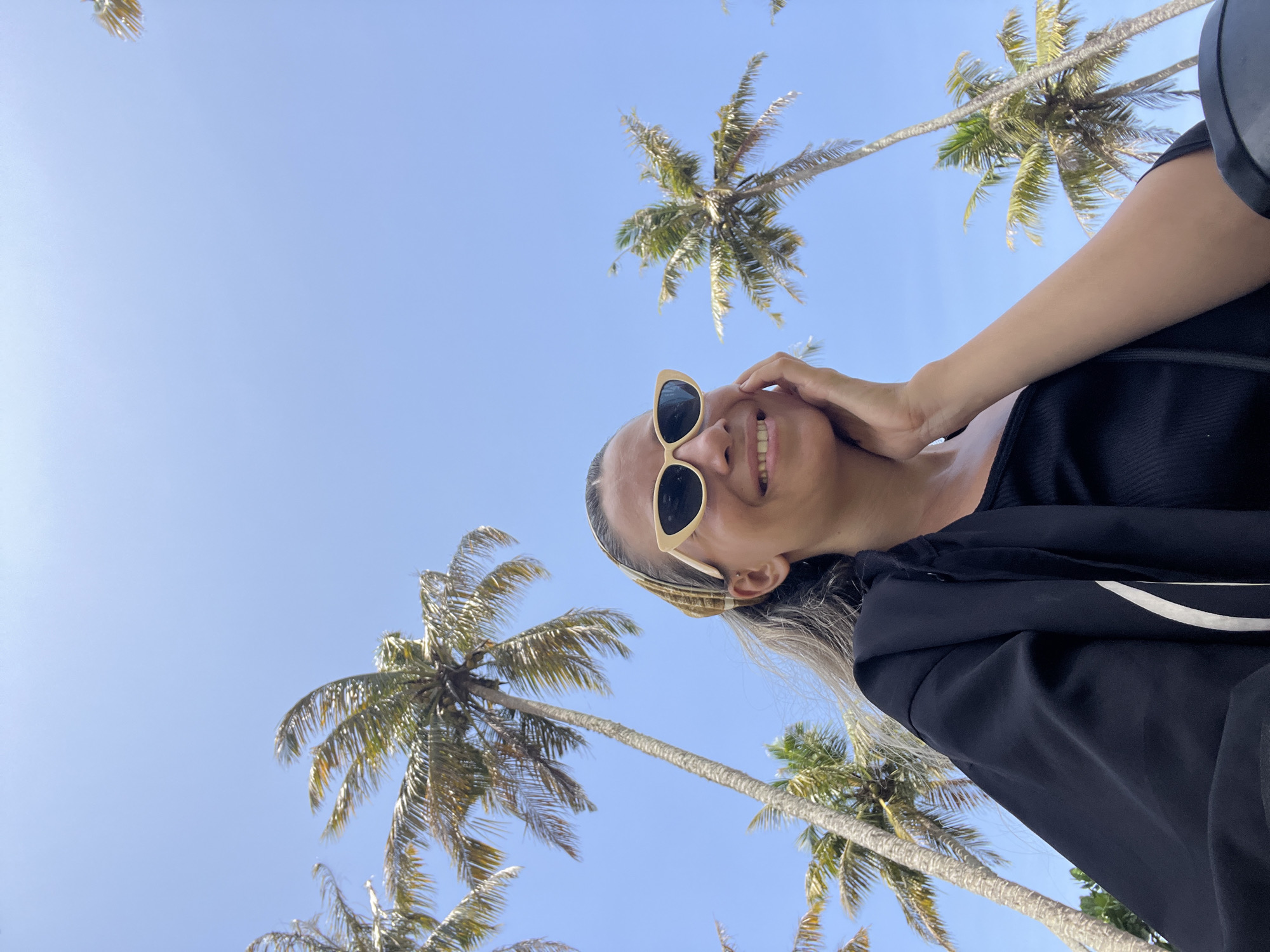 A low-angle, close-up shot captures a woman in Dondra, Sri Lanka, with a bright blue sky and several palm trees in the background. The woman is wearing cat-eye sunglasses with pale yellow frames, a black top, and a black jacket. She has a light-colored headscarf around her hair and is resting her right hand on her cheek, smiling. Her left arm is visible, and she appears to be outdoors.