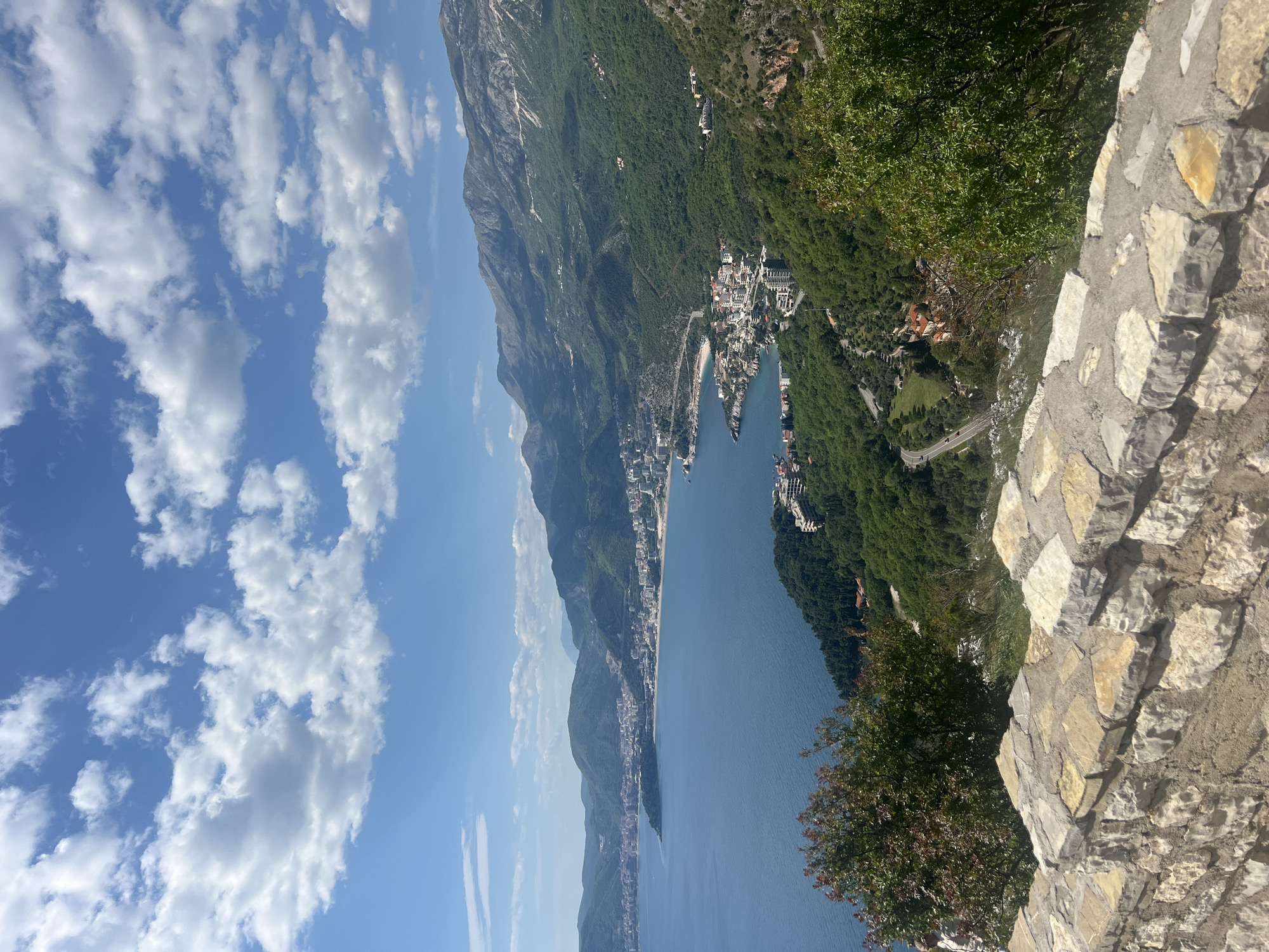 The image presents a stunning, expansive landscape view of a coastal region, likely Blizikuće, Montenegro, captured from a high vantage point. The scene is dominated by a clear blue sky adorned with scattered white cumulus clouds, suggesting a bright and sunny day.
In the foreground, a sturdy stone wall, composed of irregular, light-colored stones, frames the right side of the image, implying a viewing platform or ancient structure. Lush green foliage and trees extend from behind this wall and down the slopes, indicating dense vegetation on the mountainside.
The mid-ground showcases a tranquil, deep blue body of water, characteristic of the Adriatic Sea, curving gently along a coastline. Along this coastline, a series of charming towns and settlements are visible, with numerous buildings featuring light-colored facades. A winding road can be seen connecting these coastal communities.
Towering majestically in the background are imposing mountains, heavily covered in verdant green forests, showcasing their rugged contours. The mountains descend gradually towards the sea, creating a dramatic and picturesque backdrop for the coastal settlements. The overall impression is one of serene natural beauty, combining the grandeur of mountains, the tranquility of the sea, and the charm of human habitation. No specific activities or people are distinctly visible, and there is no discernible text in the image.