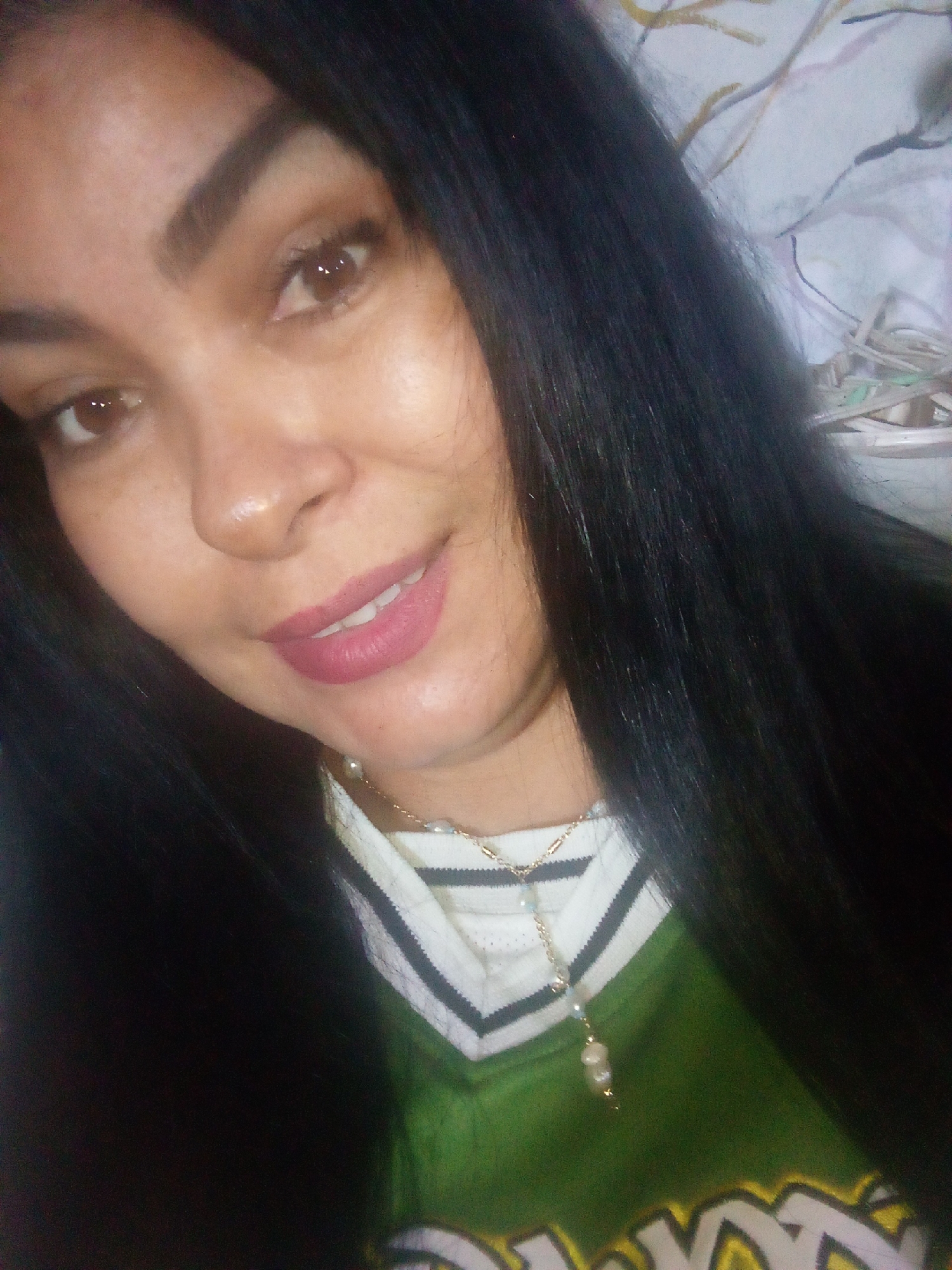 A woman with long, dark hair is posing for a selfie. She has brown eyes, arched eyebrows, and is wearing pink lipstick. She is wearing a green and white sports jersey and a gold necklace with pearl pendants. The background appears to be a cluttered room. There is no indication of a location in Juan Griego, Venezuela in the image.