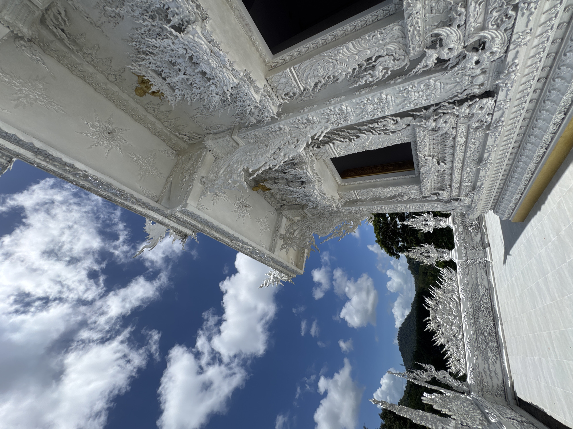 The image shows the exterior of a white, ornate temple building against a bright blue sky with scattered white clouds. The temple features intricate white plasterwork with decorative motifs, including what appear to be stylized flames and mythical creatures. The architecture has sharp, angular lines and detailed ornamentation. In the background, lush green hills and trees are visible, suggesting a natural, possibly mountainous, environment. The foreground shows a white tiled patio. The scene is likely outdoors and the lighting suggests it is daytime. The building itself is the primary subject, with the sky and surrounding landscape forming the environment. There are no people visible in the image. The location is identified as Thep Krasatti, Thailand.