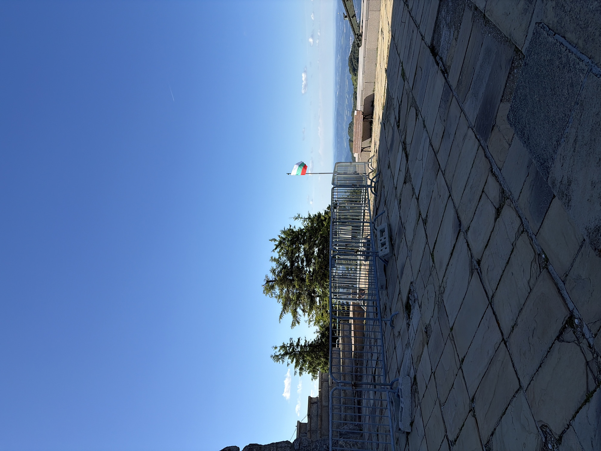 This image captures a sunny day at the Monument to the Freedom of Bulgaria, also known as the Shipka Monument, located in the city of Shipka, Bulgaria. The foreground features a stone-tiled plaza, with some tiles cracked or missing, suggesting wear and tear. A temporary metal barrier fence is set up, with concrete blocks used as stabilizers. Behind the fence, a stone monument or structure is visible on the left, with tiered steps leading up.
In the middle ground, several tall coniferous trees stand against a clear blue sky, with a few scattered white clouds. A Bulgarian flag, featuring white, green, and red horizontal stripes, is prominently displayed on a flagpole, waving gently. Further back, a wooden bench is placed on a slightly elevated platform. The background reveals a vast, rolling landscape of hills and valleys, suggesting a mountainous or elevated location. The overall impression is one of a scenic overlook and a historical monument.