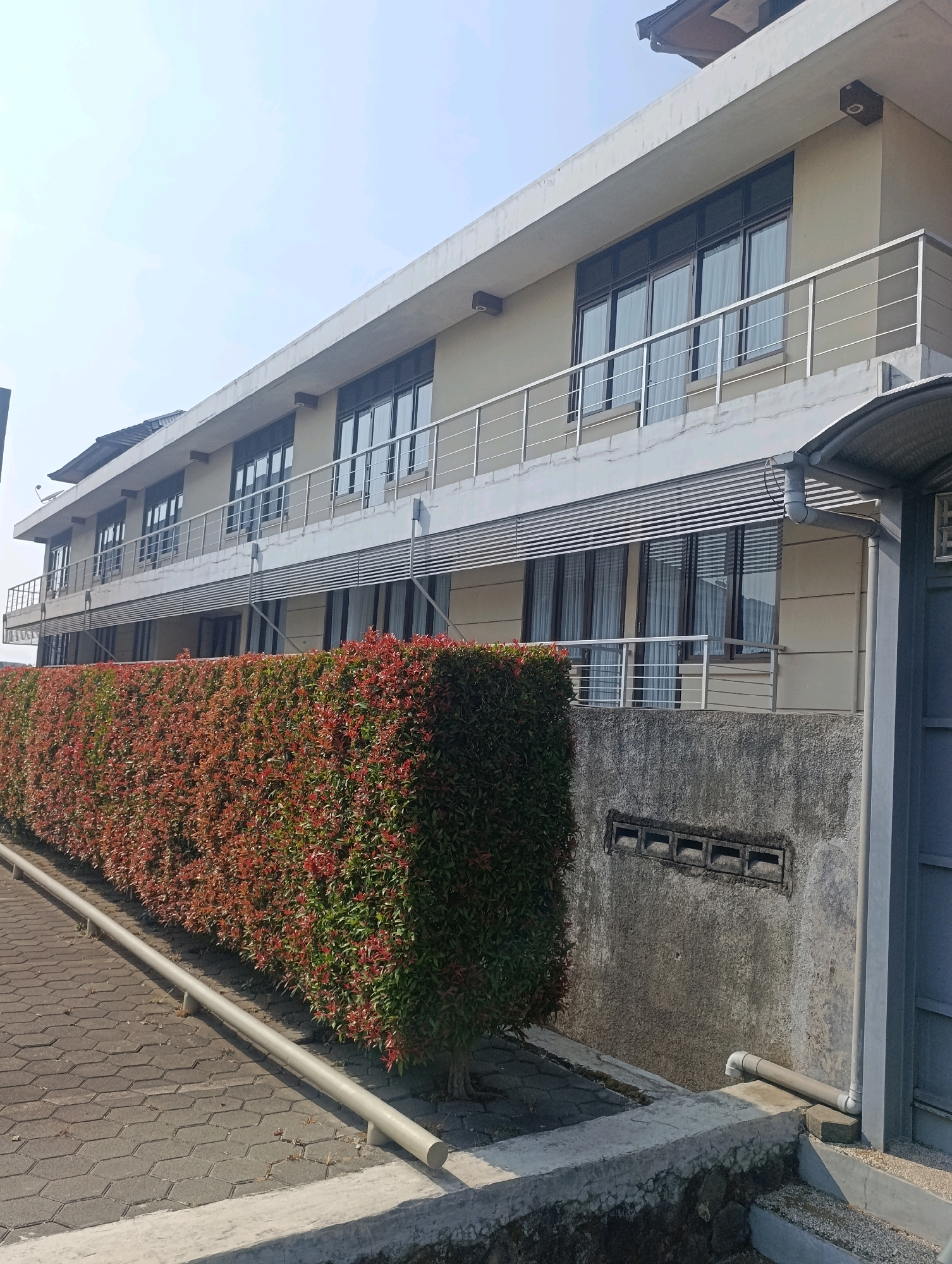 The image shows a three-story building with a beige exterior and a white overhang. The building has multiple windows with dark frames and white curtains visible inside. A silver railing runs along the balconies of the building. In front of the building, a dense hedge with red and green leaves is visible. The ground in front of the hedge is paved with hexagonal gray tiles, and a silver railing runs along the edge of the paved area. A concrete wall with rectangular vents is located behind the hedge. A gray pipe is attached to the wall and runs down to the ground. The scene appears to be a residential or commercial property in a city.