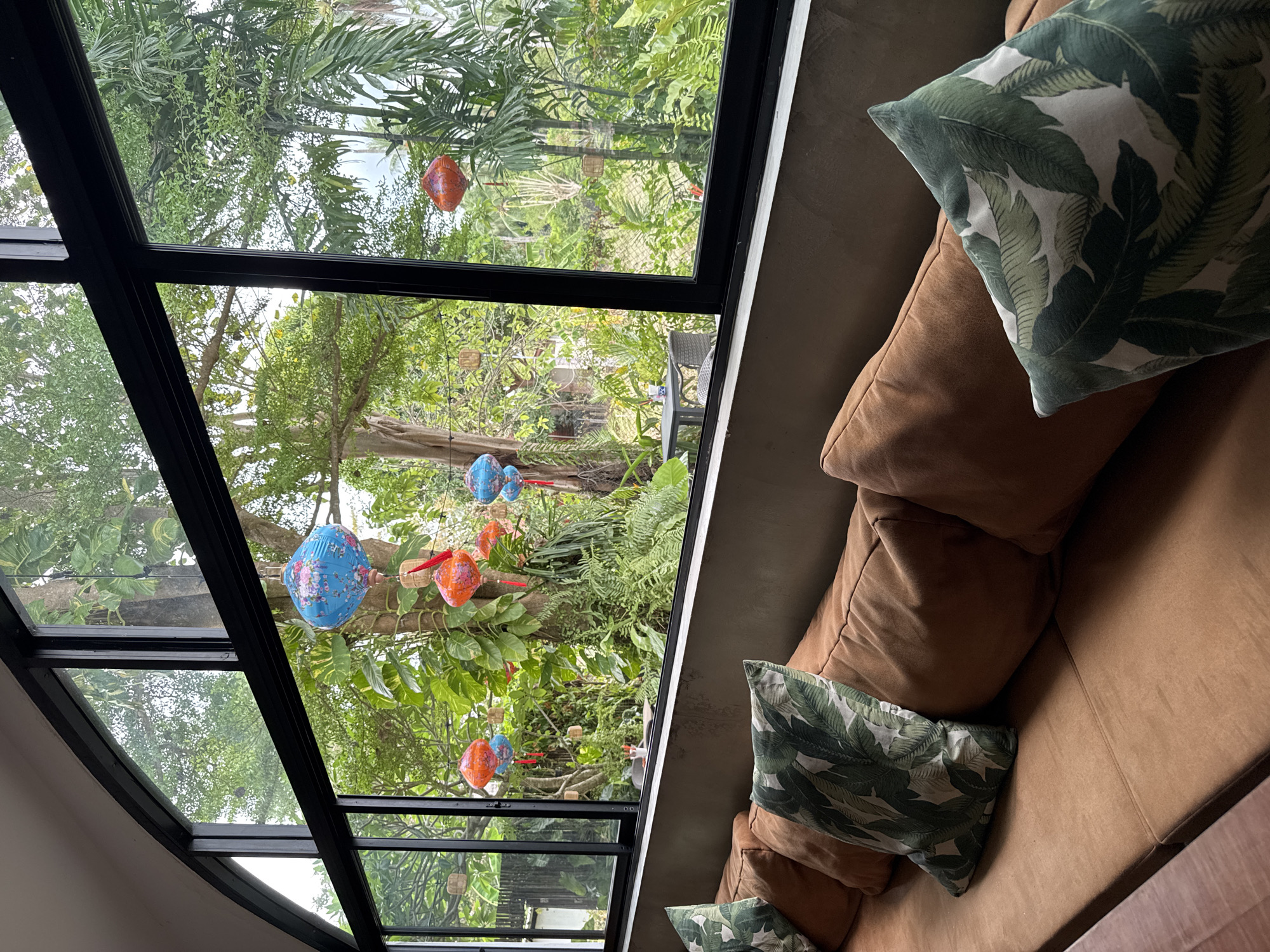 The image shows the interior of a room in Baan Mae Nam, Thailand, with a view through a large, multi-paned window of a lush, tropical garden.
The window frames are black and divide the view into multiple rectangular sections. The garden is dense with various trees and plants. Decorative lanterns of different colors (blue, orange) are suspended from the branches of a tree in the garden.
Inside, a tan or light brown sofa is visible, with several cushions. Some of the cushions are a solid tan/brown color, while others have a white and green tropical leaf pattern. The walls of the room appear to be a concrete color.