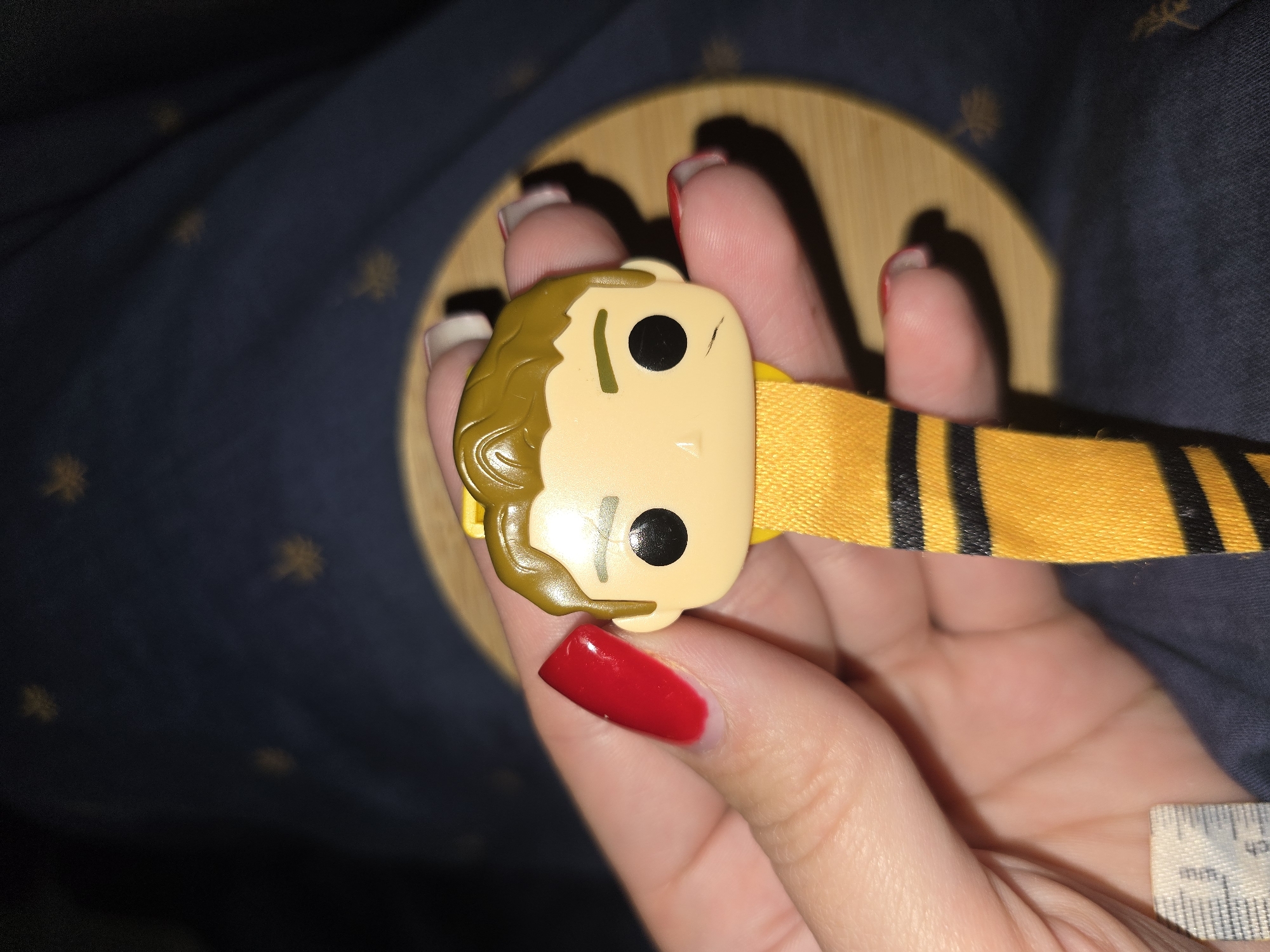 A close-up, eye-level shot shows a person's hands holding a Funko Pop keychain featuring a character with light brown hair, black eyes, and a yellow ribbon with black stripes attached. The background is a dark blue fabric with faint yellow patterns. The scene is indoors, with no discernible city or specific location information.