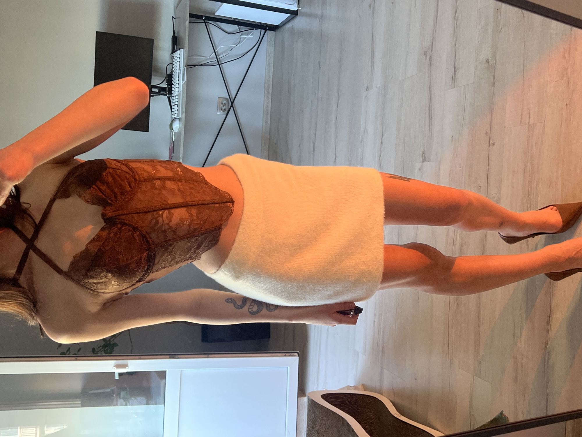 This media file features a single individual in what appears to be a home or personal space. The subject is standing, with the camera angled from a low perspective, capturing them from the mid-torso down to their feet. The person is wearing a brown lace corset and a fluffy white mini skirt. They have a snake tattoo visible on their forearm. They are also wearing brown high-heeled shoes.
The background shows a wooden floor and a section of a wall. In the distance, a desk with a computer monitor and keyboard is partially visible, suggesting an office or study area within the setting. There's also a hint of a large window with blinds in the lower part of the frame, and a glimpse of some furniture or shelving units. The lighting in the scene is warm, possibly from artificial light sources, creating shadows on the floor.
The image is characterized by its intimate and personal nature. The subject's pose and attire suggest a focus on fashion or a personal moment captured at home. The time of day is not explicitly indicated, but the warm lighting could imply an evening setting. No weather conditions are discernible. There is no visible text in the image. The overall mood is somewhat posed and artistic, with the warm lighting and the subject's posture contributing to a sophisticated aesthetic.