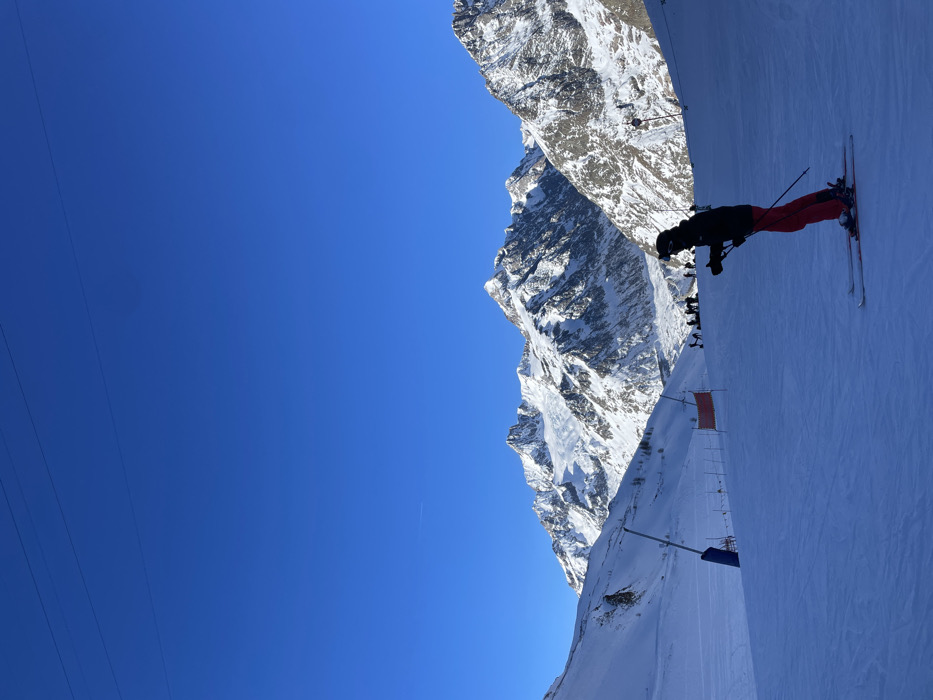The image displays a bright, sunny day on a snow-covered mountain slope in Courmayeur, Italy. In the foreground, positioned on the right, a skier is visible, dressed in a black jacket, red pants, and a helmet, standing with skis and poles on the groomed snow. The vast majority of the left side of the frame is filled with a clear, deep blue sky, indicating excellent weather.
In the background, rugged, snow-capped mountains rise majestically, showcasing their rocky and snowy peaks and slopes under the bright sunlight. Along the ski slope, particularly further down, other smaller figures of people or skiers are discernible, along with what appears to be orange safety netting or flags. Thin lines, likely ski lift cables or power lines, cut across the upper left section of the sky. The scene depicts a typical winter sports activity with perfect visibility and conditions, likely during mid-day or early afternoon given the strong sunlight and clear shadows. No legible text is visible in the image.