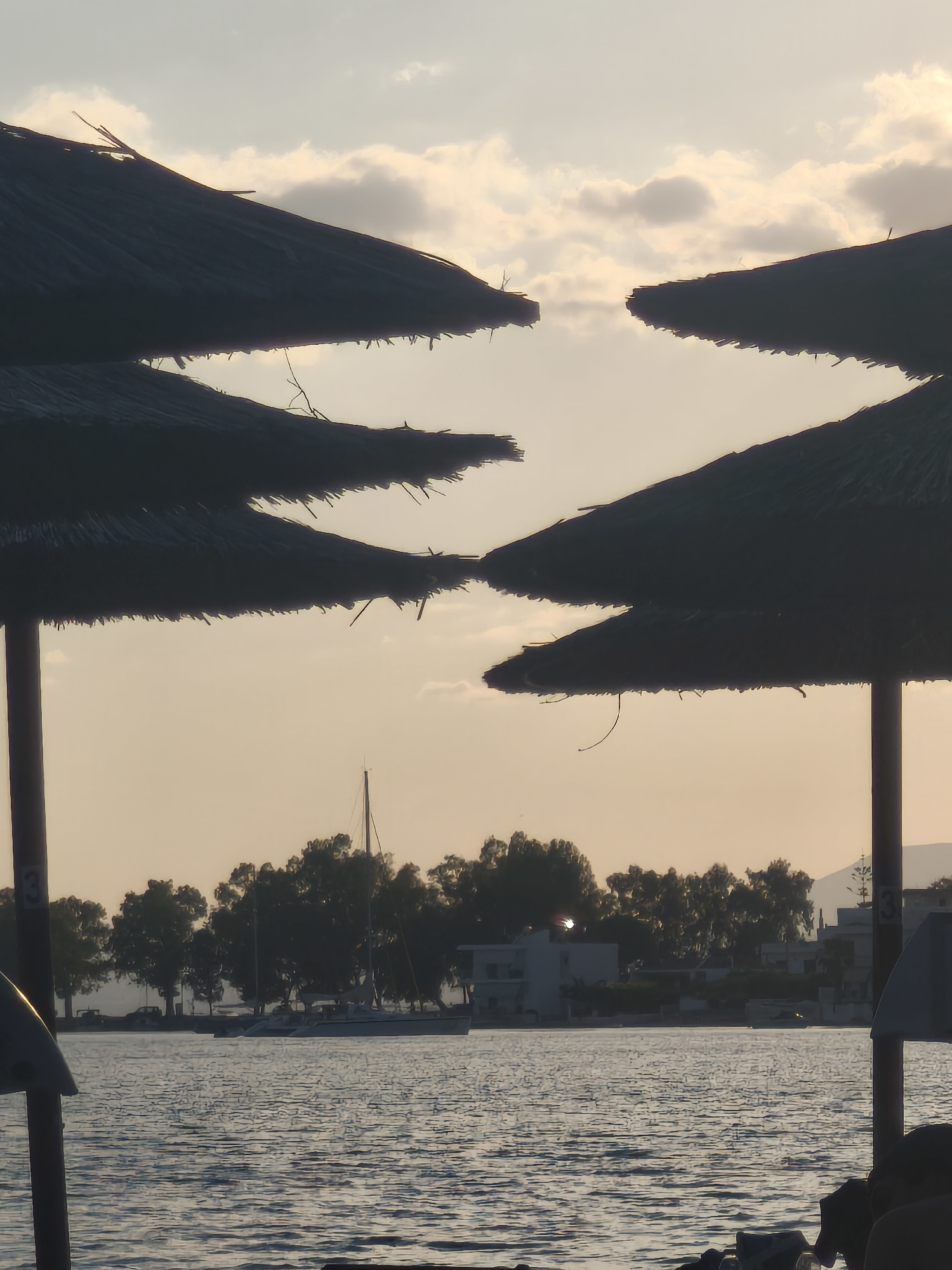 The image depicts a serene waterfront scene in Eretria, Greece, likely at sunset or sunrise, given the soft, warm lighting and diffused sky. In the foreground, the silhouettes of two thatched-roof umbrellas dominate the frame, their rustic texture a stark contrast to the calm water below. One umbrella is positioned on the left, its broad canopy casting a shadow, while another, larger umbrella, is on the right, partially obscuring the view. Between these umbrellas, the tranquil sea stretches towards a distant shoreline.
The middle ground reveals a picturesque coastline lined with lush green trees, interspersed with buildings that suggest a coastal town or resort. The distinct masts of several sailboats are visible against the backdrop of the trees and sky, hinting at maritime activity. The water itself is gently rippled, reflecting the subdued light of the sky.
In the distance, a faint mountain range is visible, adding depth to the landscape. The sky is a canvas of soft hues, with scattered clouds that are illuminated by the low-angled sun. The overall impression is one of peace and natural beauty, characteristic of a Mediterranean coastal setting. The presence of the umbrellas suggests a beach or a waterfront promenade where visitors can relax and enjoy the view.