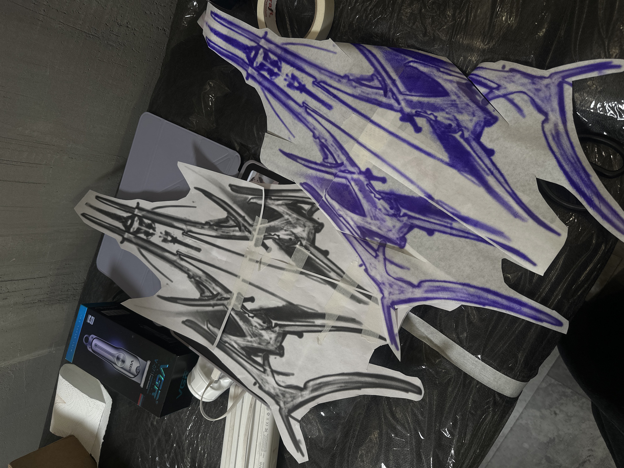 The image shows an overhead shot of tattoo-related items on a dark surface, possibly covered with plastic wrap. Two tattoo stencils, one in black and one in blue, are prominently displayed. A light blue case, a VGR hair trimmer box, a roll of tape, and a small pile of white napkins are also present. Cables are present near the trimmer box. The location is not identifiable as Moscow, Russia, or any other specific location.
